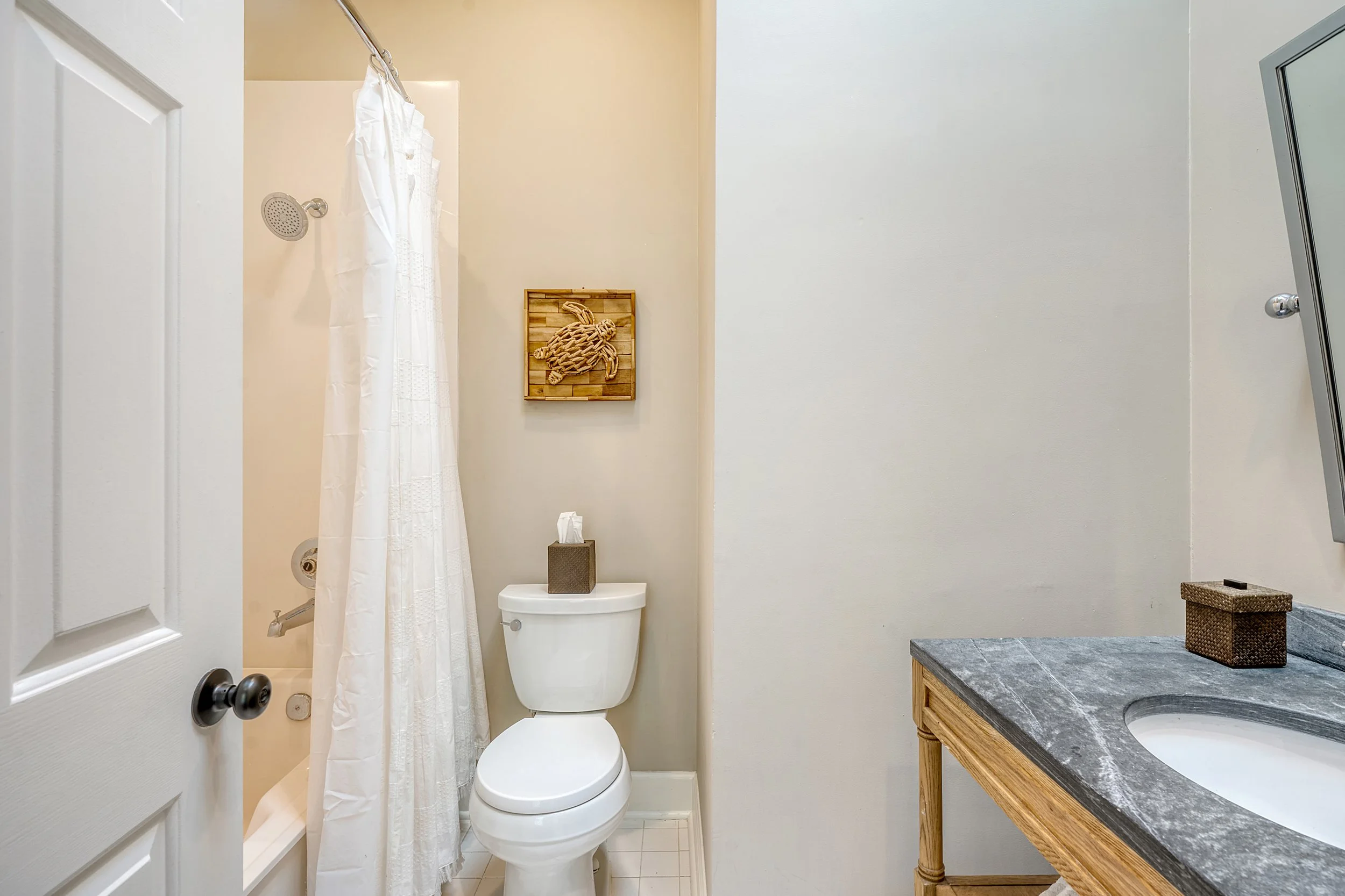 Small bathroom with a bathtub and shower on the left, a toilet in the center, and a wooden vanity with a gray countertop and sink on the right. A tissue box is on the toilet tank, and another tissue box is on the vanity. There is a decorative wooden 