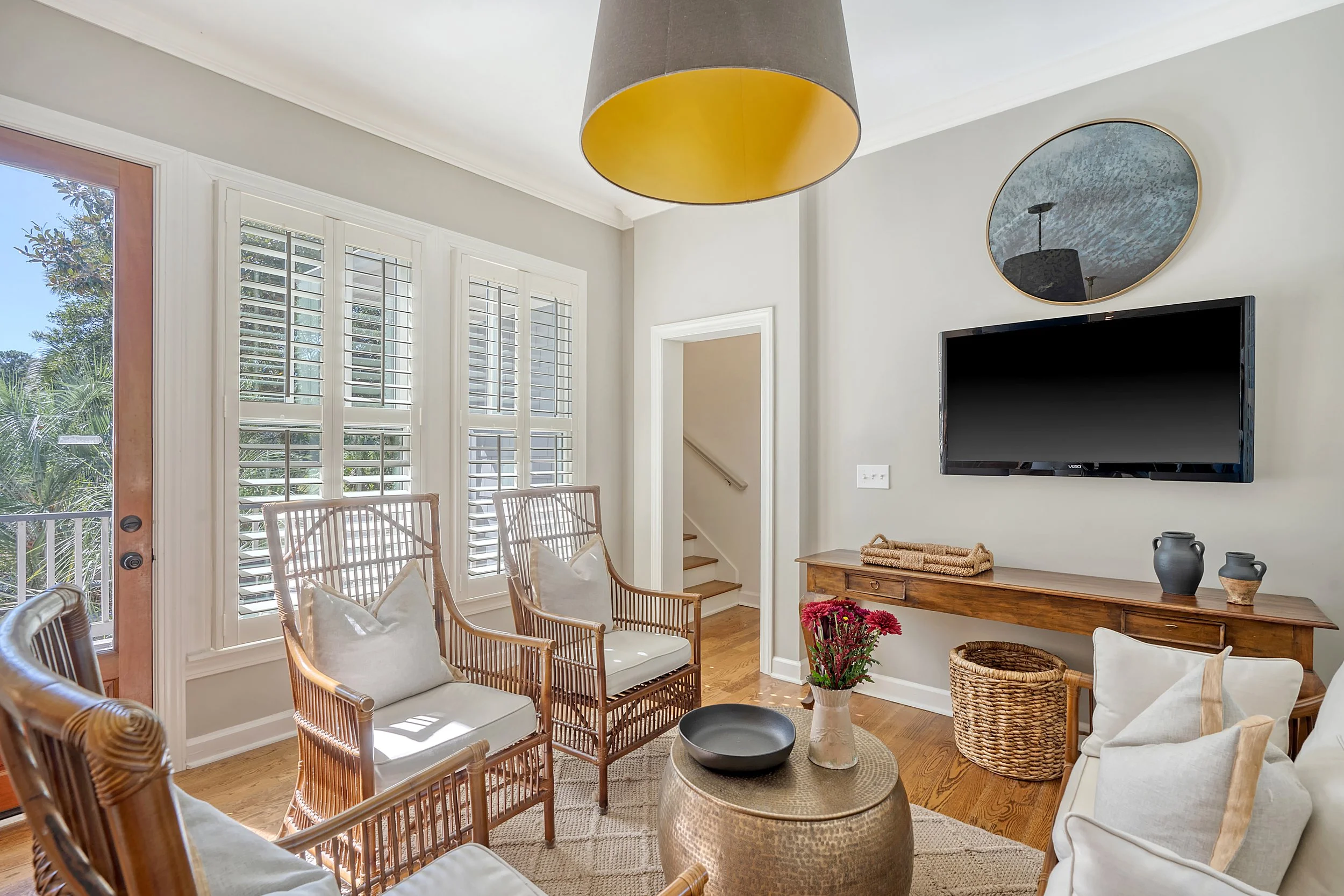 Living room with large window, wooden chairs with white cushions, a wooden console table, a flat-screen TV, round mirror, hanging ceiling light, and decorative vases and flowers.