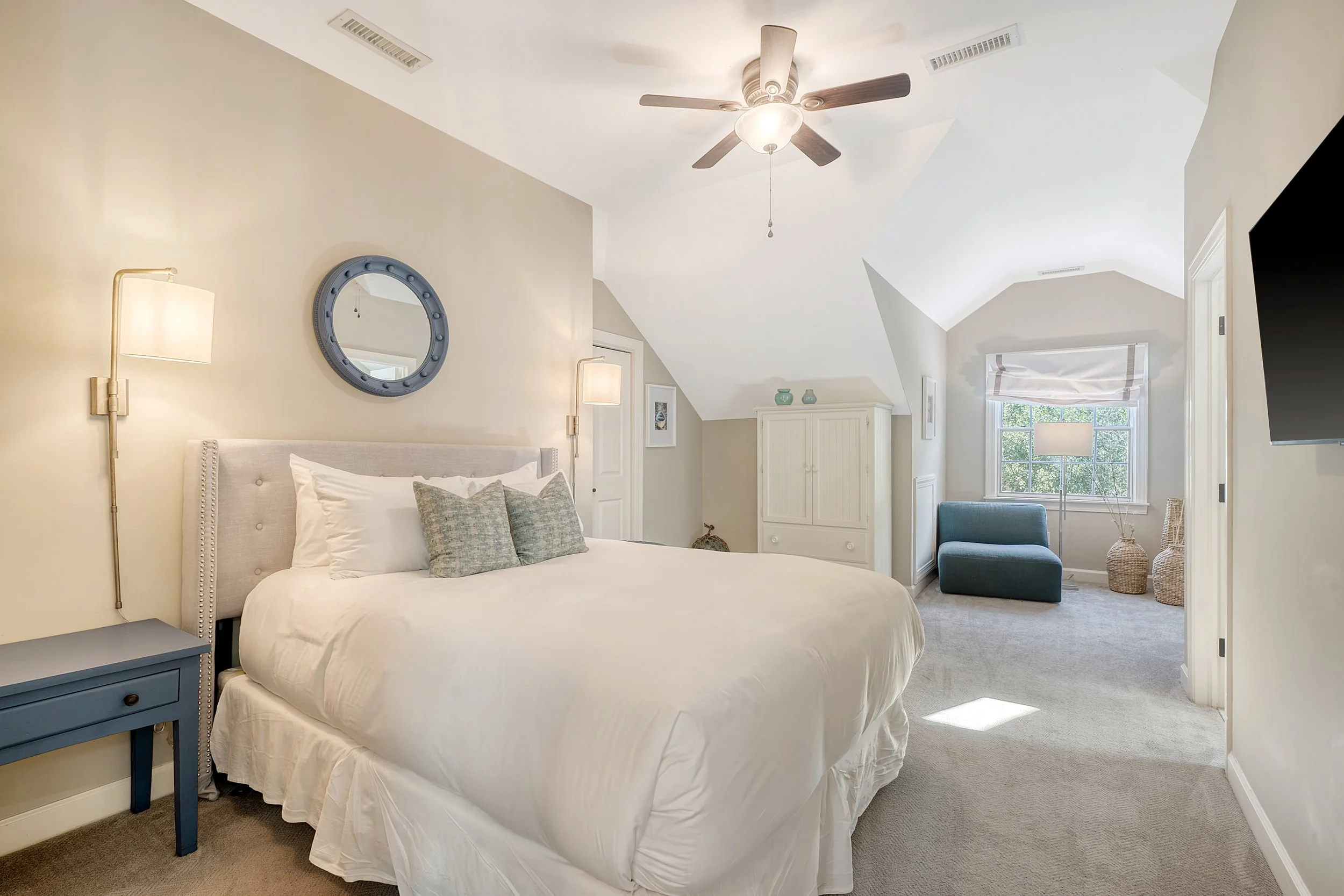 A bedroom with white walls, a white bed with pillows, and a blue bedside table. There are two wall-mounted lamps on either side of the bed, a circular mirror above the headboard, a ceiling fan overhead, and a window with a white blind letting in natu