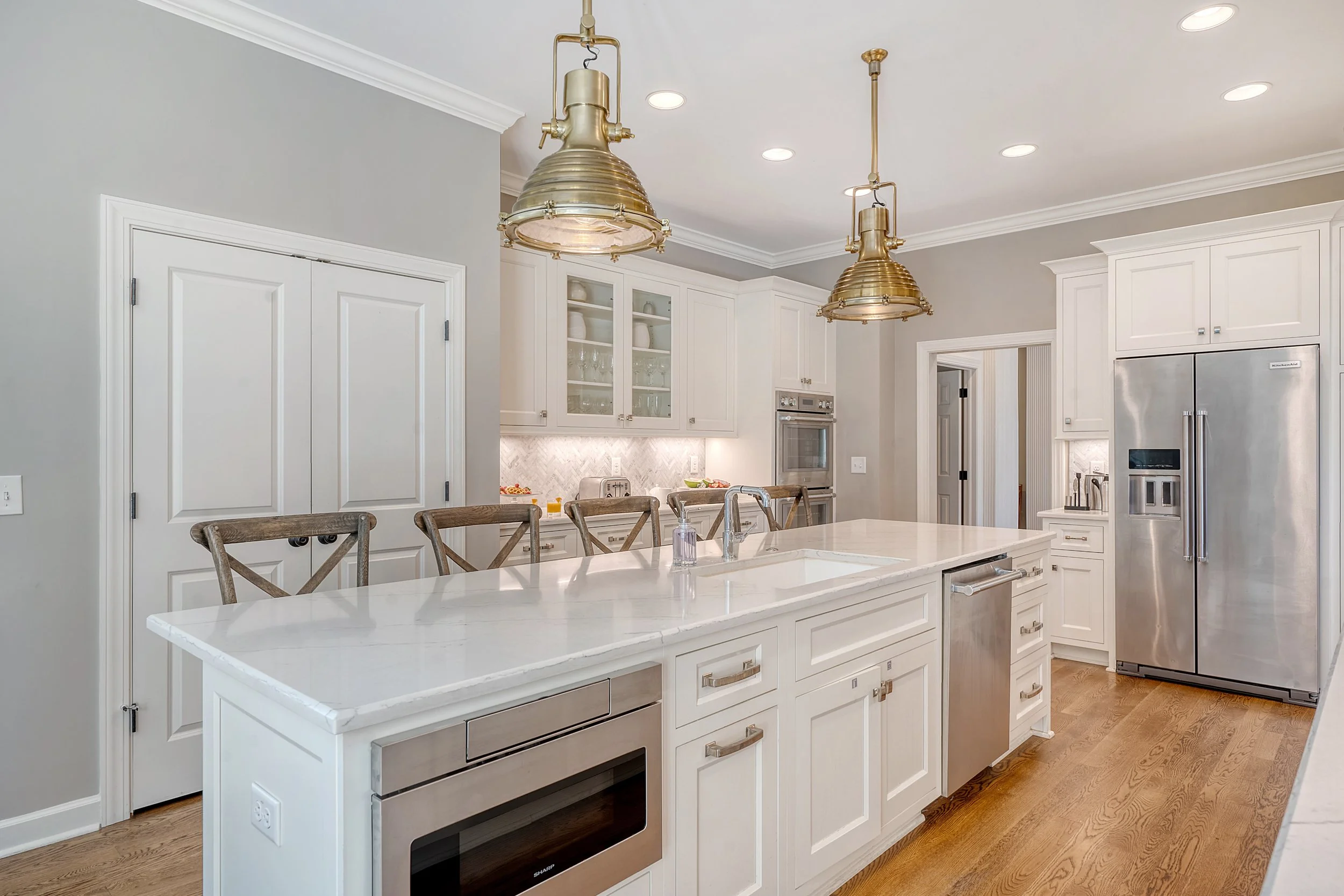 Modern kitchen with white cabinets, marble island, stainless steel refrigerator, built-in oven, and two brass pendant lights hanging from the ceiling.