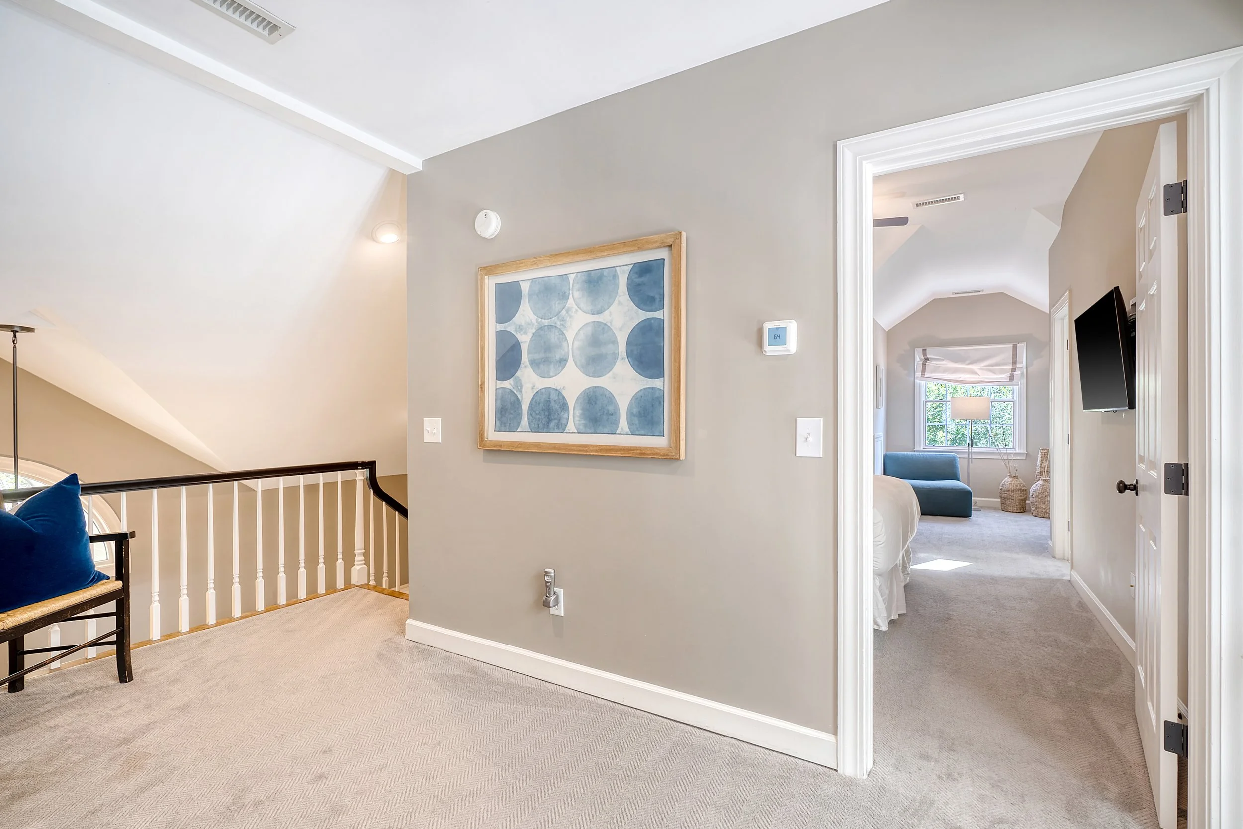 View of a house interior with a hallway leading to a bedroom and a staircase on the left side. The hallway has beige walls and carpet flooring, with a framed blue circular artwork on the wall and a thermostat.
