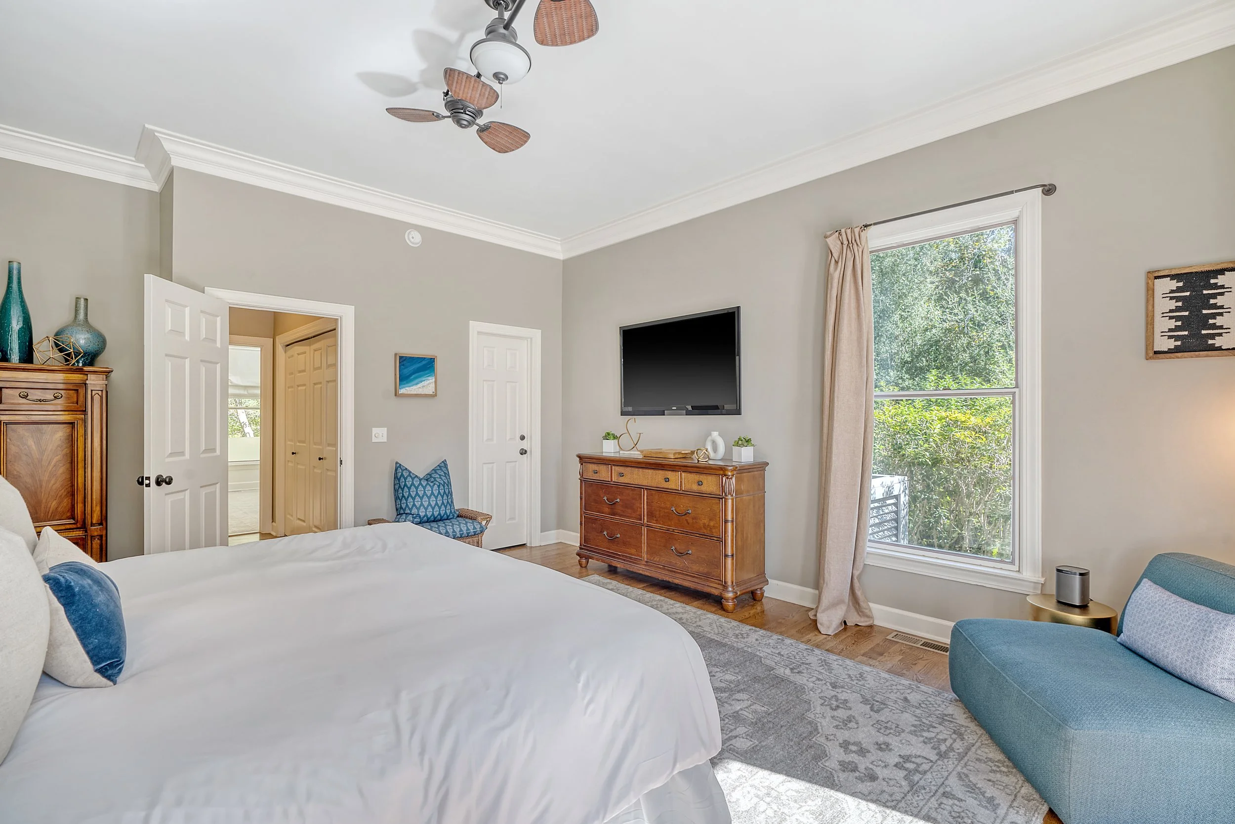A bedroom with a large bed, wooden furniture, a ceiling fan, and a window showing outdoor greenery.