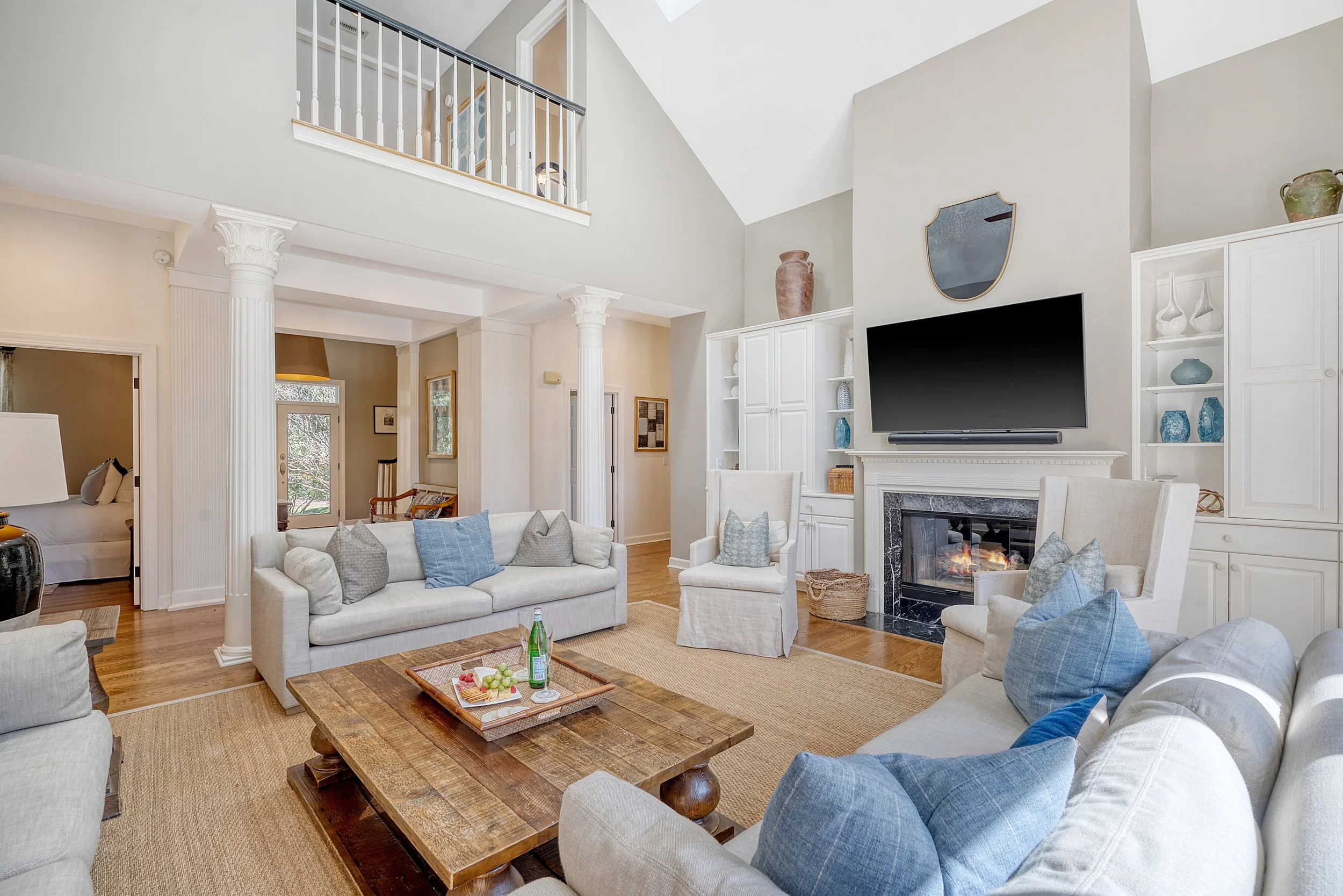Spacious living room with white sofas with blue and gray pillows, a wooden coffee table, a fireplace, a flat-screen TV, built-in shelves with decorative items, and a second-floor balcony overlooking the room.