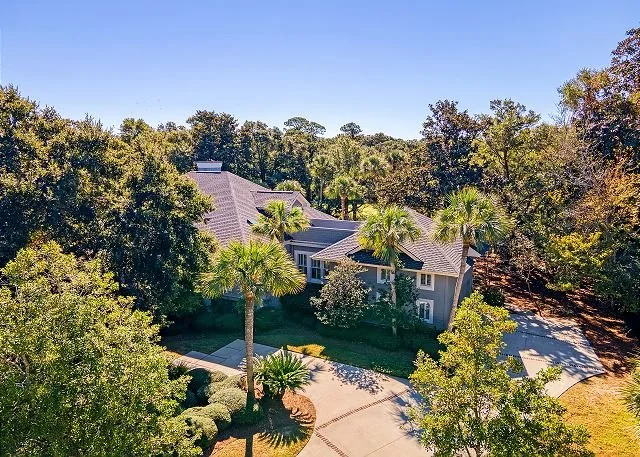 A large house surrounded by lush trees and a well-maintained yard with palm trees. A driveway curves toward the house on a sunny day.