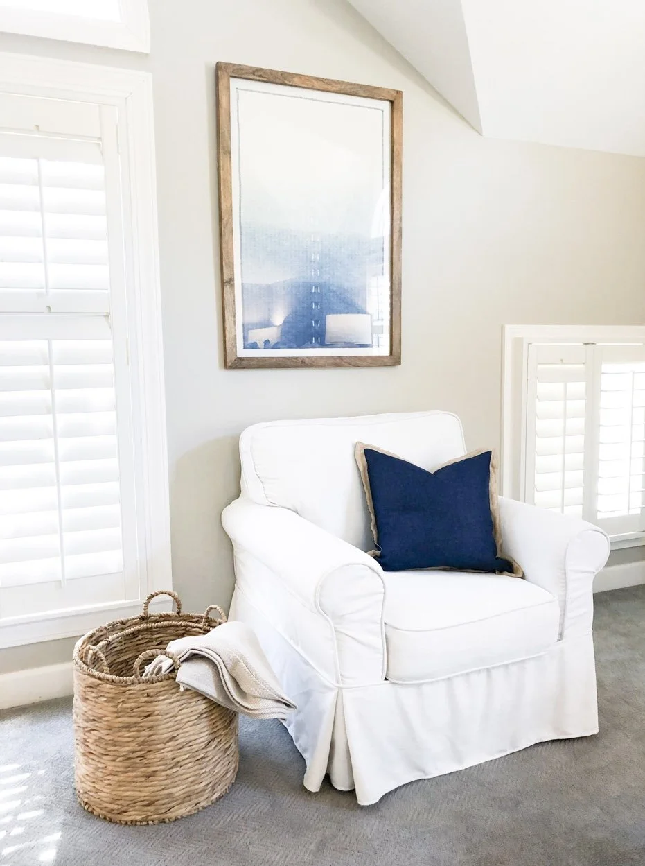 A cozy corner with a white armchair, a navy blue pillow, a woven wicker basket with a towel, and a large picture frame on the wall, in a softly lit room with white shutters.