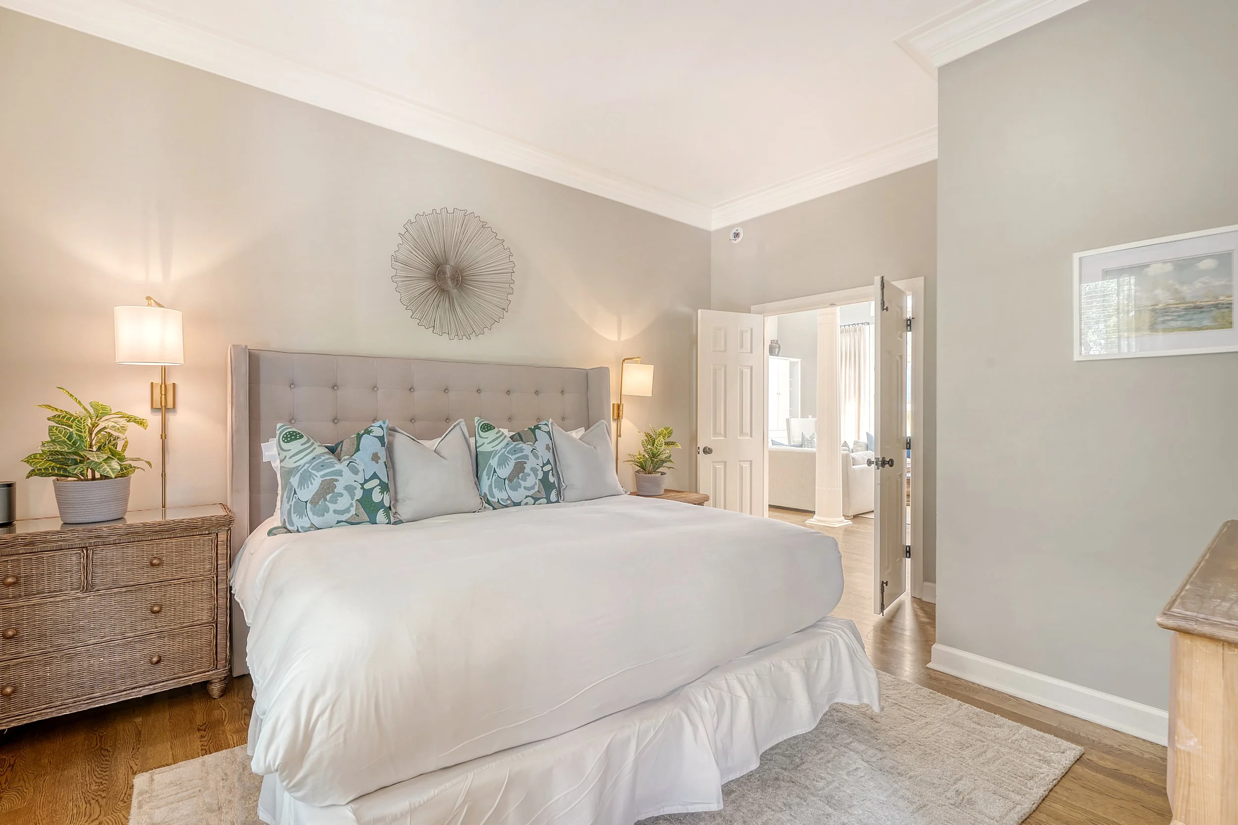 A bedroom with a large bed featuring a beige upholstered headboard, white bedding, and decorative pillows. There are nightstands with lamps and potted plants on both sides of the bed. An open door reveals another room with a sitting area and large wi