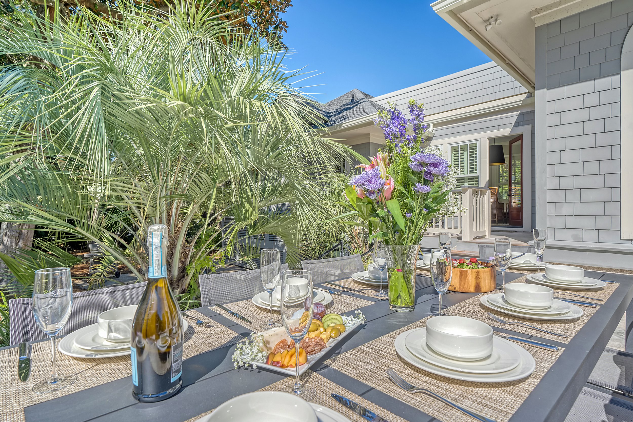 Outdoor dining table set for a meal, with a vase of purple flowers, glasses, plates, and cutlery on a dark table with woven placemats, in a backyard with lush green palm trees and a house with a porch in the background under a clear blue sky.