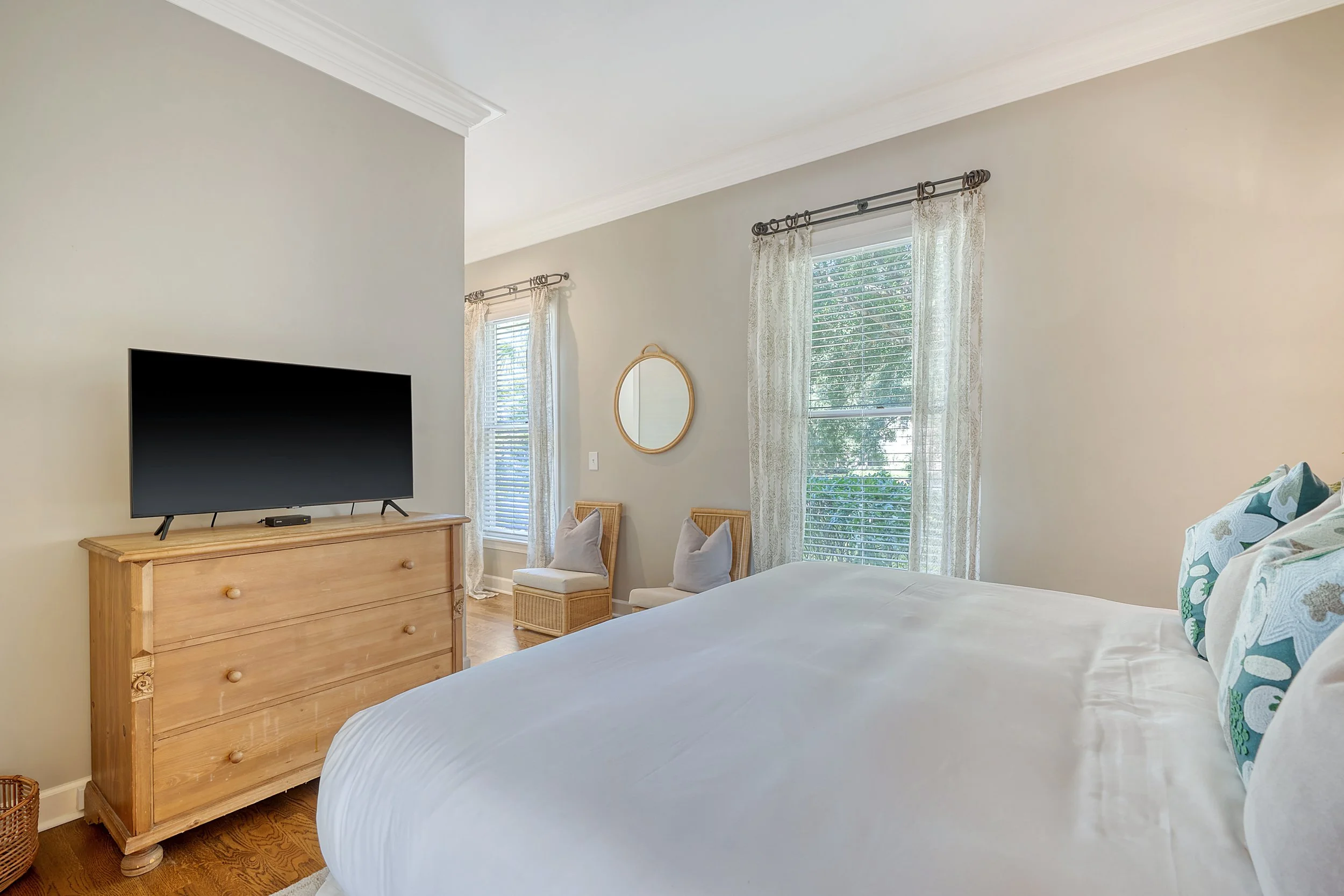 Bright bedroom with large bed and decorative pillows, wooden dresser with TV, and windows with white curtains.