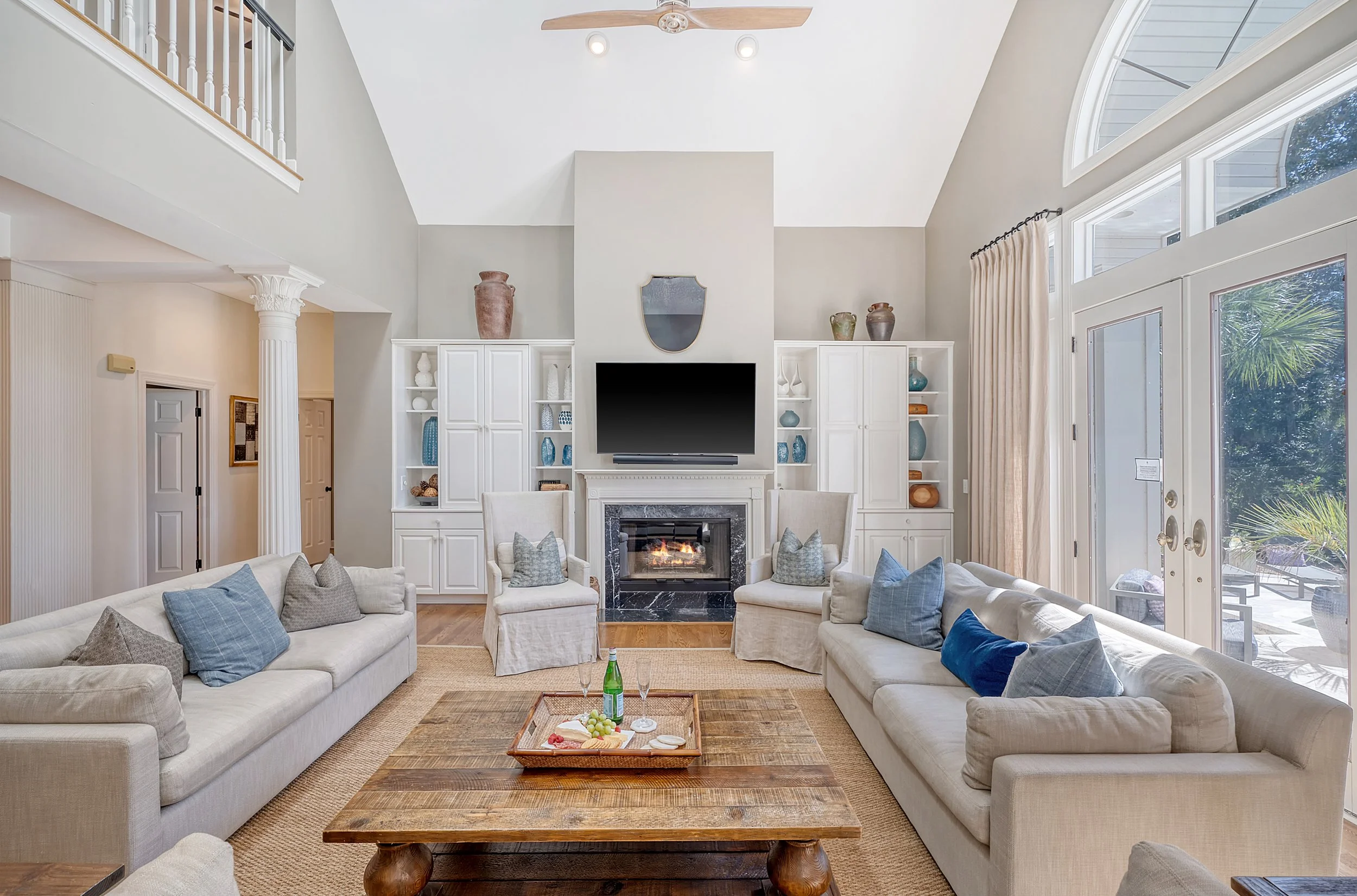 Bright living room with white sofas, a wooden coffee table, a fireplace, built-in white shelves, a large flat-screen TV, and large windows with curtains.