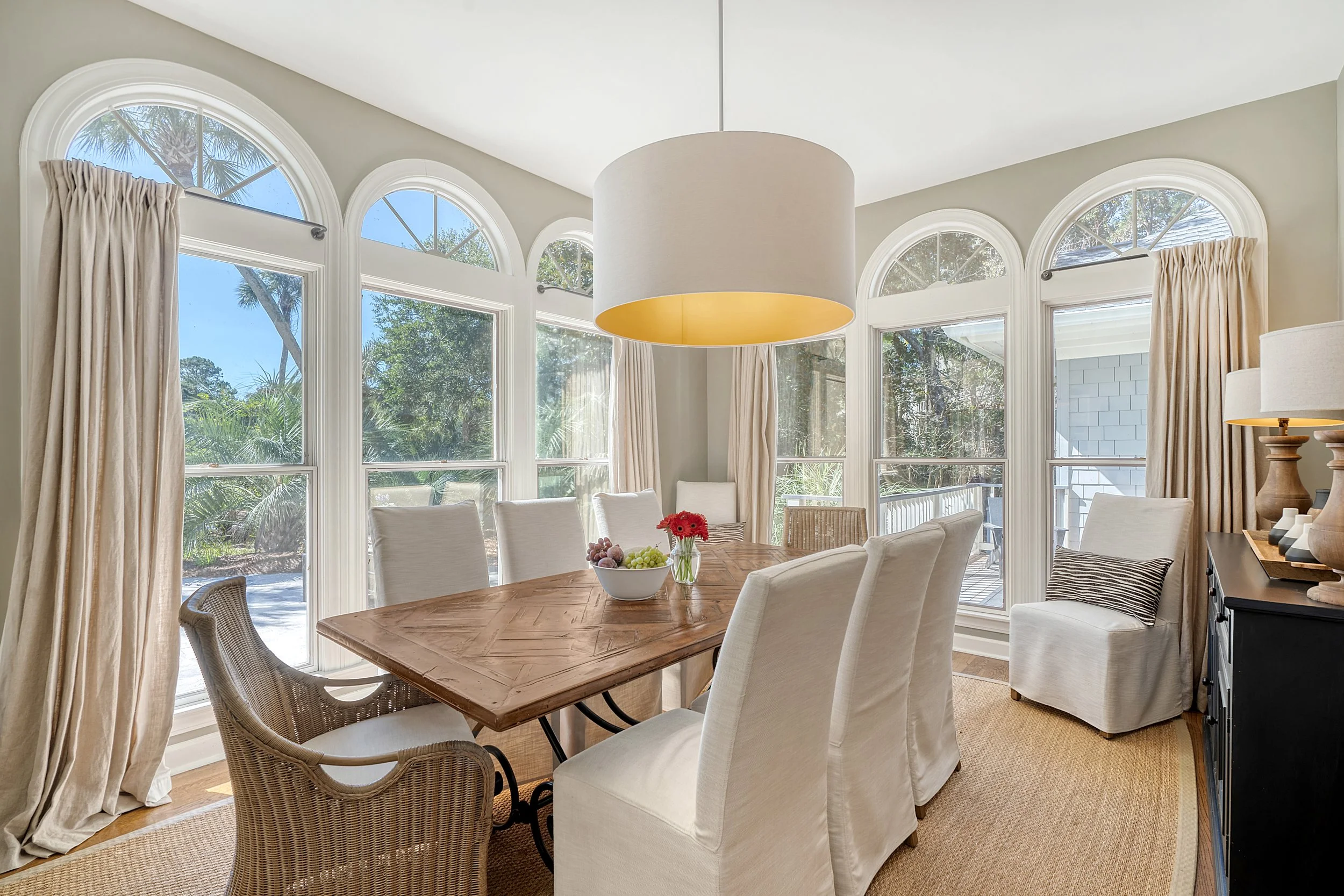 Dining room with large arched windows, tan curtains, wooden table, and chairs, with a bowl of fruit and flowers, sunlight streaming in.
