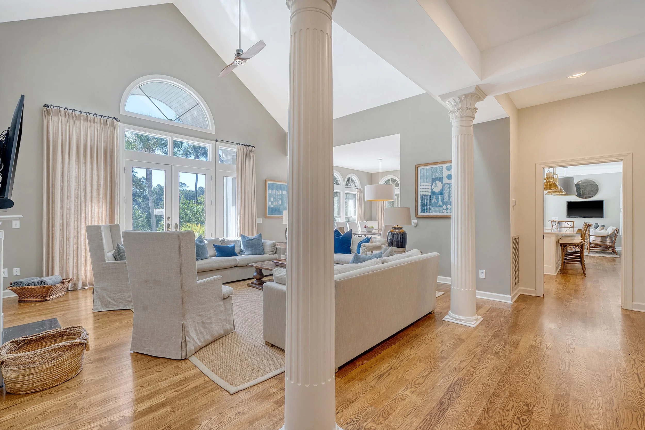 A spacious, well-lit living room with large windows, cream-colored sofas, and wooden floors, separated into two areas by white decorative columns.