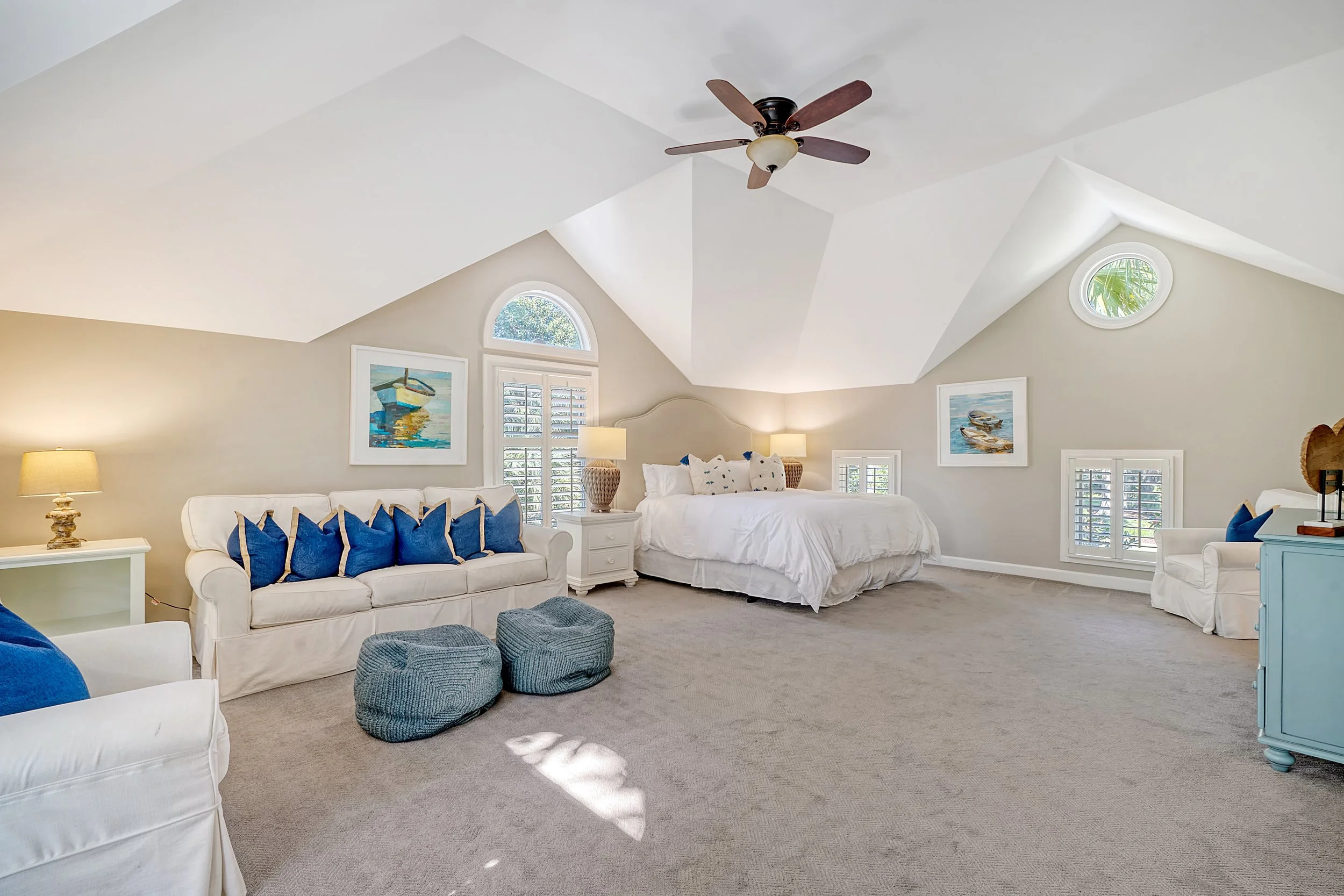 Bright bedroom with sloped ceiling, white bed with multiple pillows, white sofa with blue pillows, blue ottomans, framed artwork of boats, windows with shutters, ceiling fan.