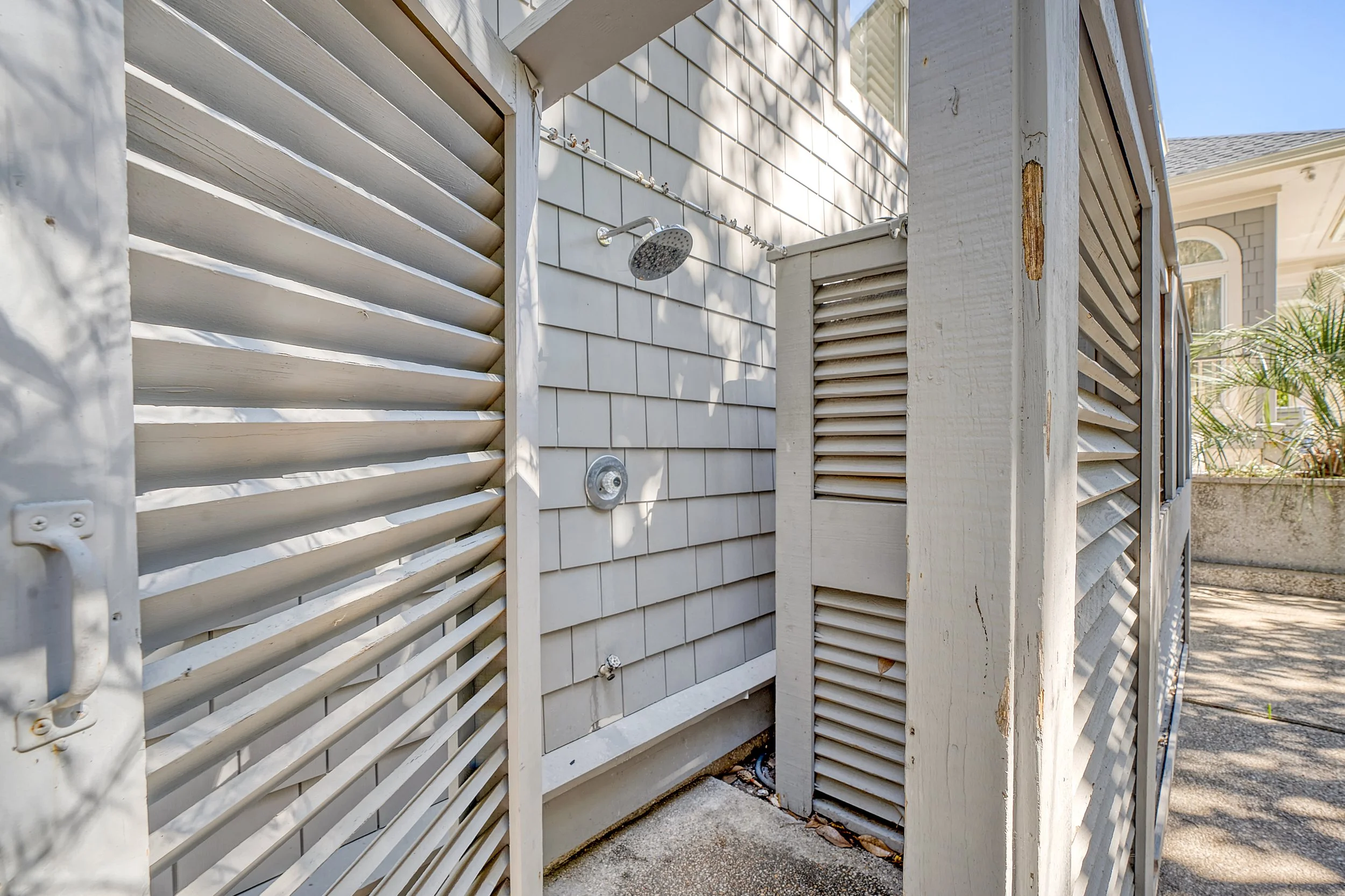 View of an outdoor shower area featuring a shower head, water control knob, white wooden shutters, and a wall with beige shingles, with sunlight and nearby plants.