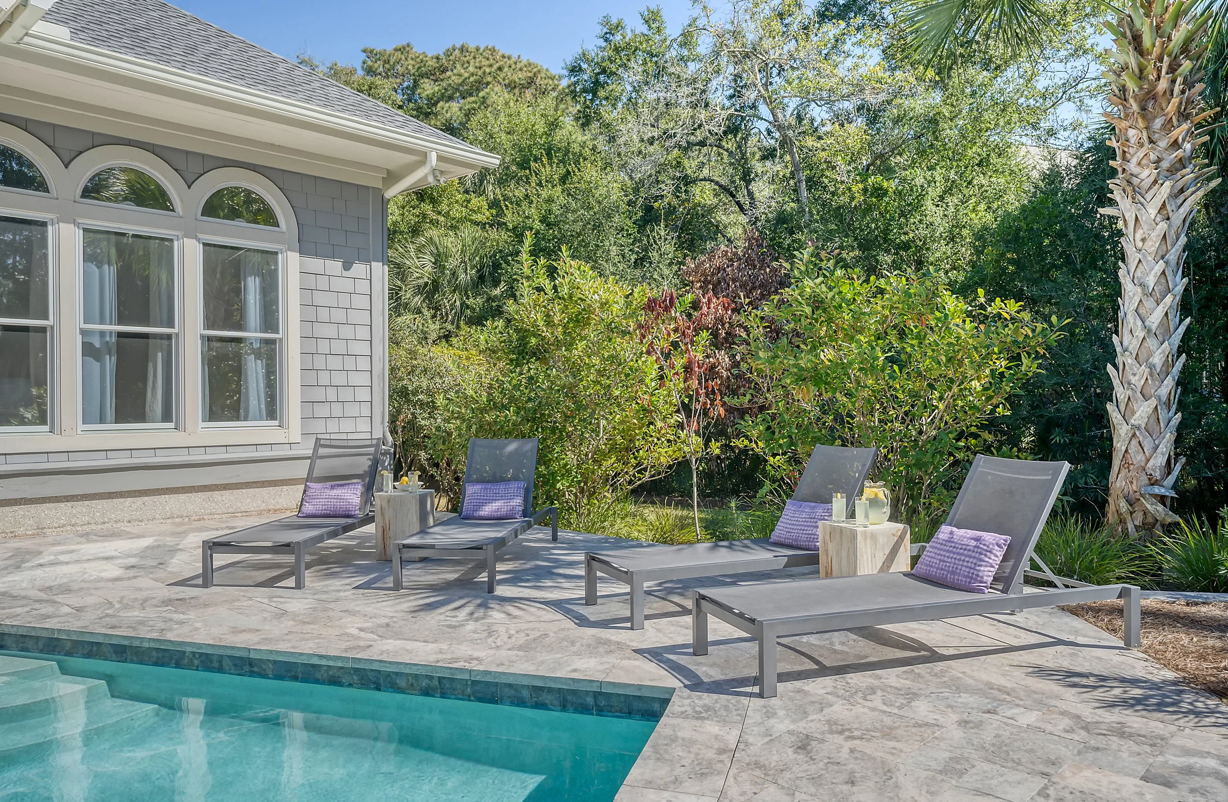 Sunbathing area with four lounge chairs with purple pillows, small side tables, and a swimming pool in the foreground. Lush green trees and bushes in the background near a house with gray siding and arched window.