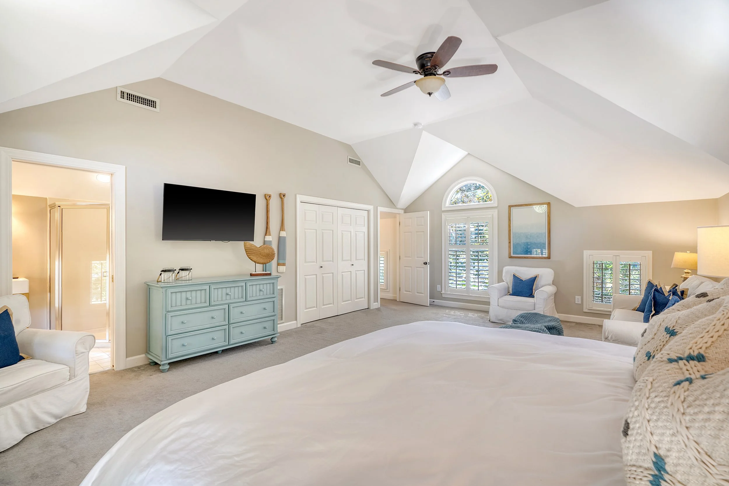 Bright bedroom with a vaulted ceiling, ceiling fan, large bed, white armchair, blue throw pillows, and a blue dresser. There are multiple windows with shutters, a mirror, and a nearby doorway to a bathroom.