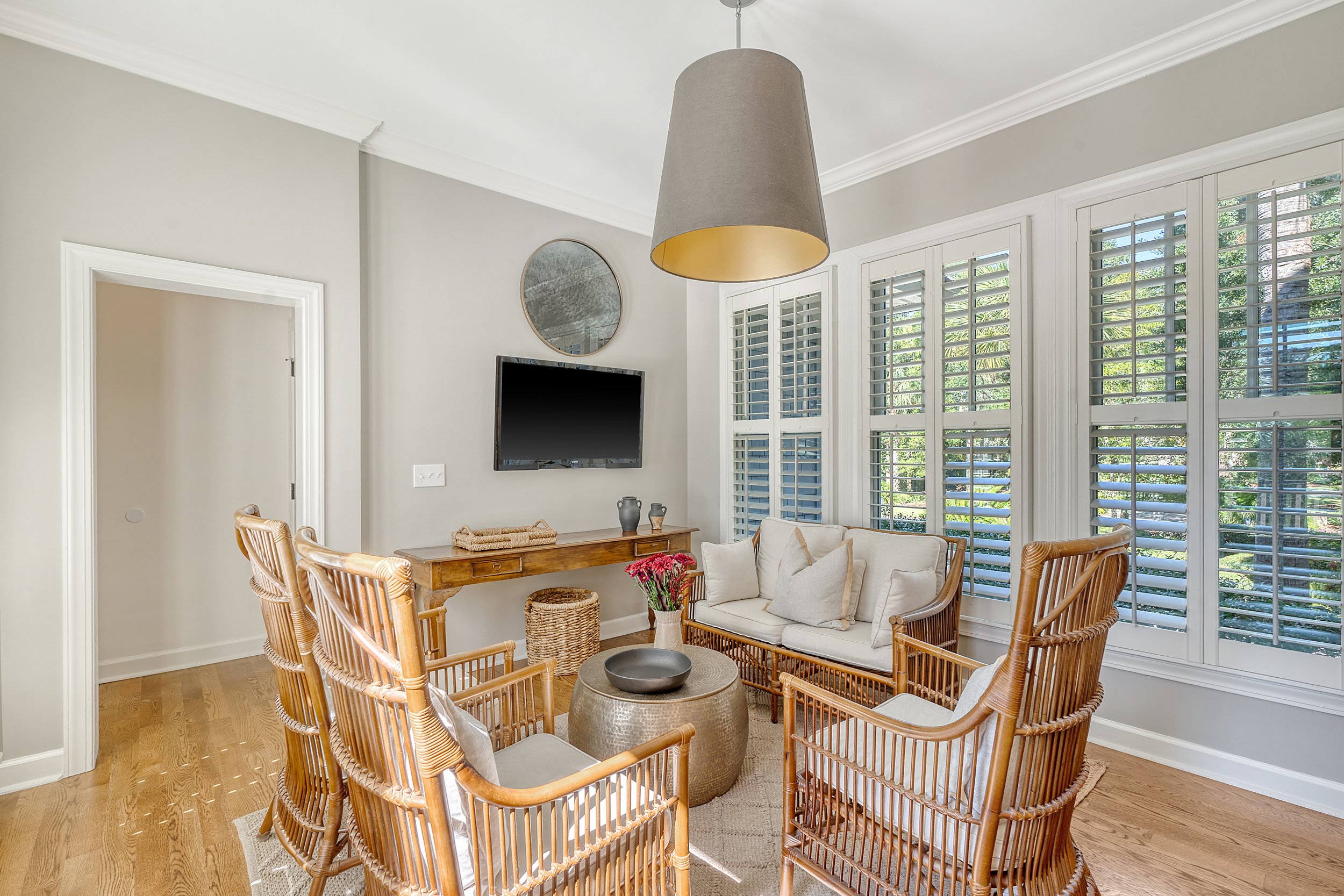 Living room with a window wall covered in white plantation shutters, a wood table, a TV mounted on the wall, and wicker chairs surrounding a small round table with a flower vase.