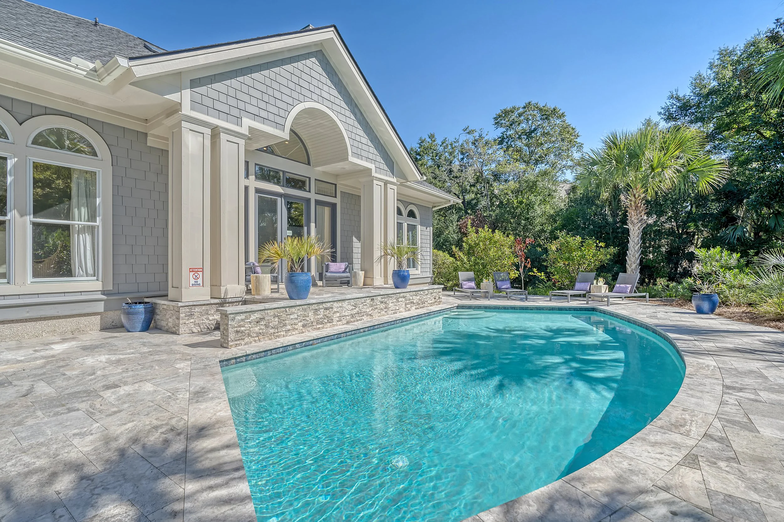 A backyard with a swimming pool, lounge chairs, potted plants, and a house with large windows and a patio under a clear blue sky.