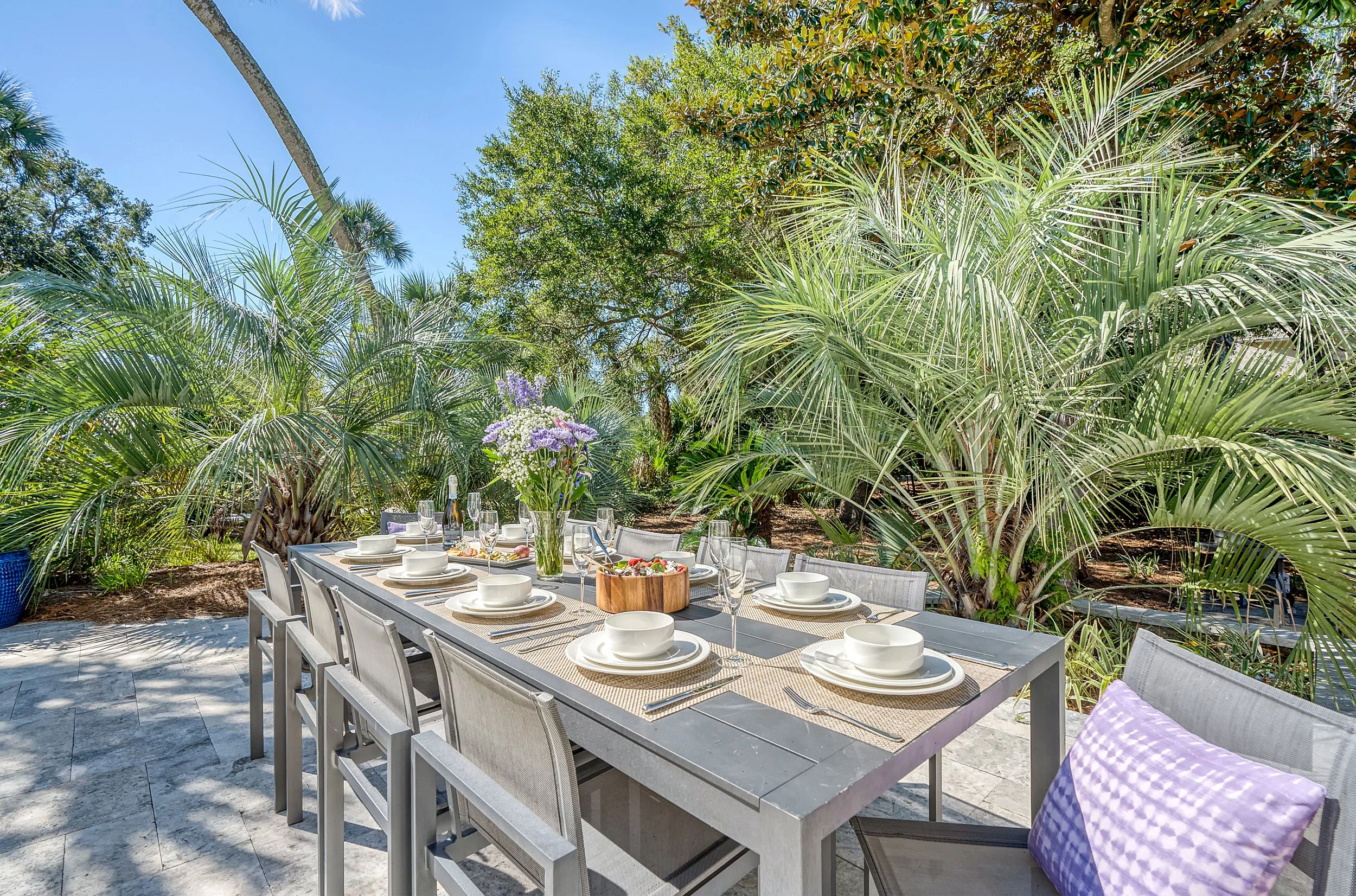 Outdoor dining table set for a meal, decorated with a centerpiece of purple and white flowers, surrounded by lush green tropical plants under a clear blue sky.