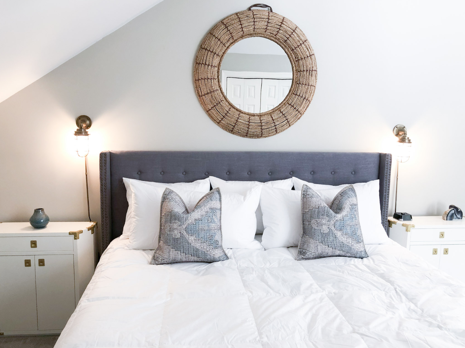 A bedroom with a bed, gray upholstered headboard, white bedding, and two decorative pillows. Two nightstands with lamps and small decor items flank the bed. A round wicker mirror hangs above the headboard.
