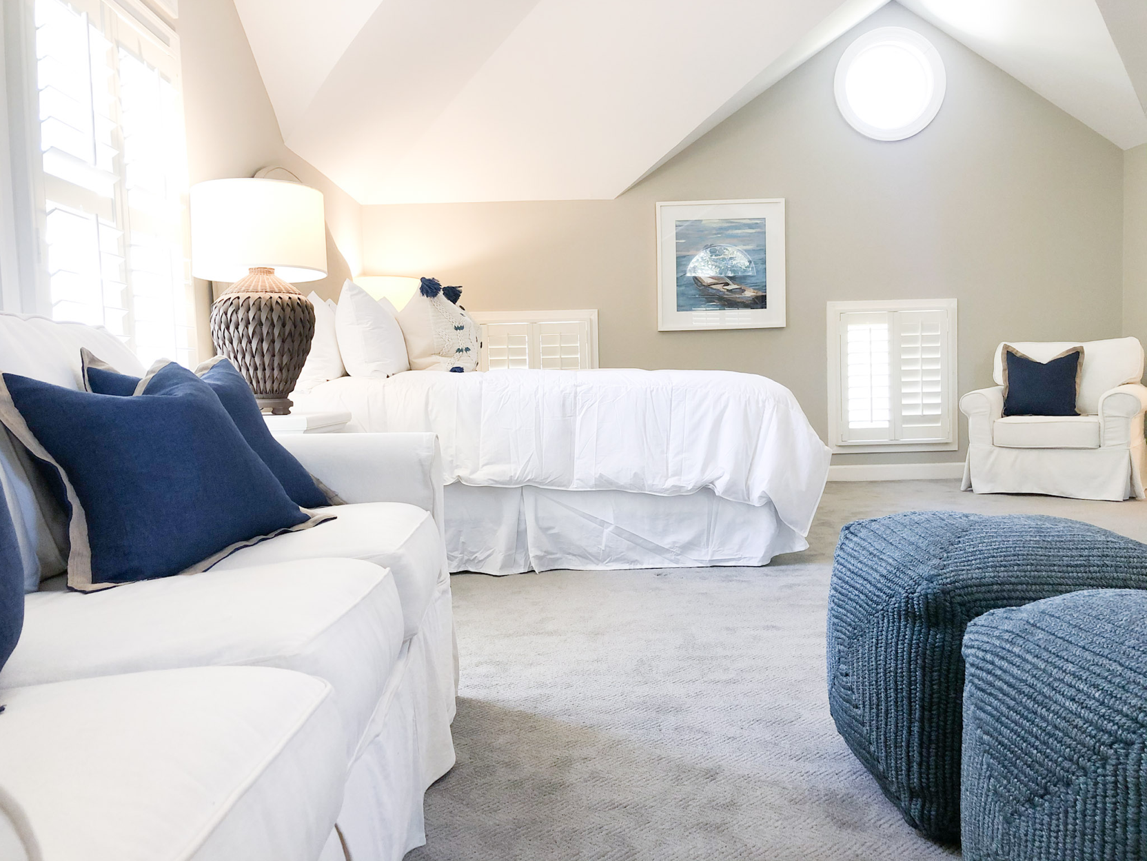 A bright bedroom with a white bed, navy and white pillows, a lamp on a side table, a white armchair with a navy pillow, another armchair in the foreground, framed artwork on the wall, and windows with white shutters.