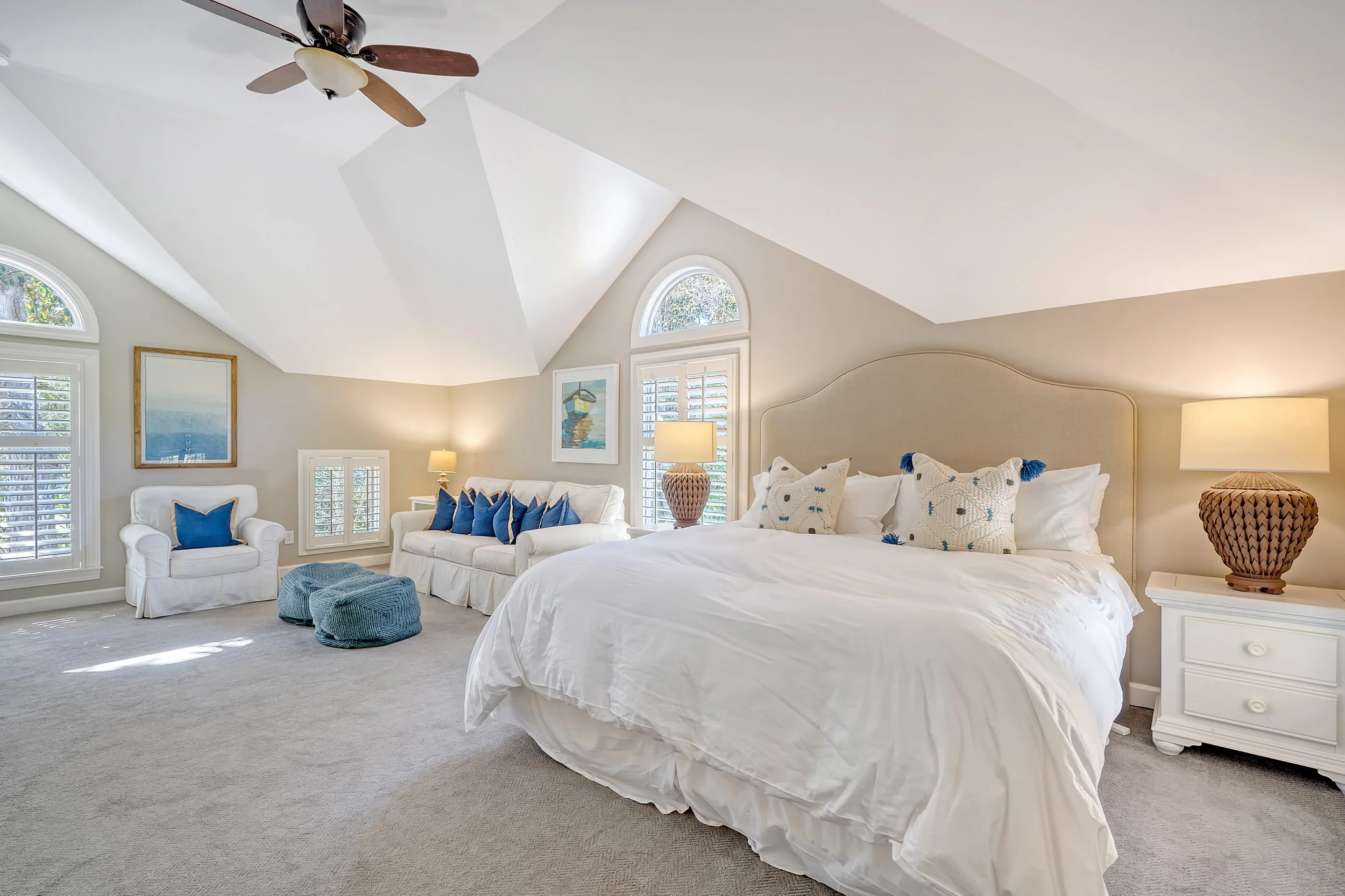 A spacious bedroom with a vaulted ceiling, white walls, and large windows with shutters. It has a white bed with decorative pillows, nightstands with lamps, a white armchair with a blue pillow, and a white sofa with blue pillows. There are two blue p