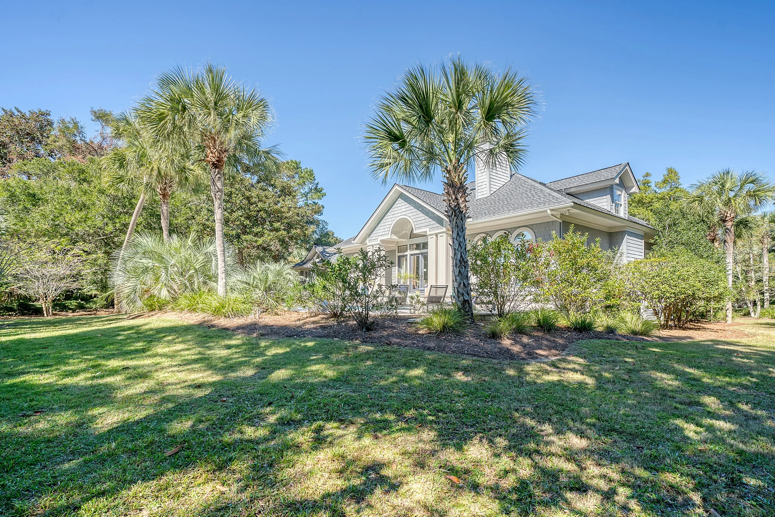 A house with grey siding and a porch is surrounded by palm trees and greenery in a sunny yard.