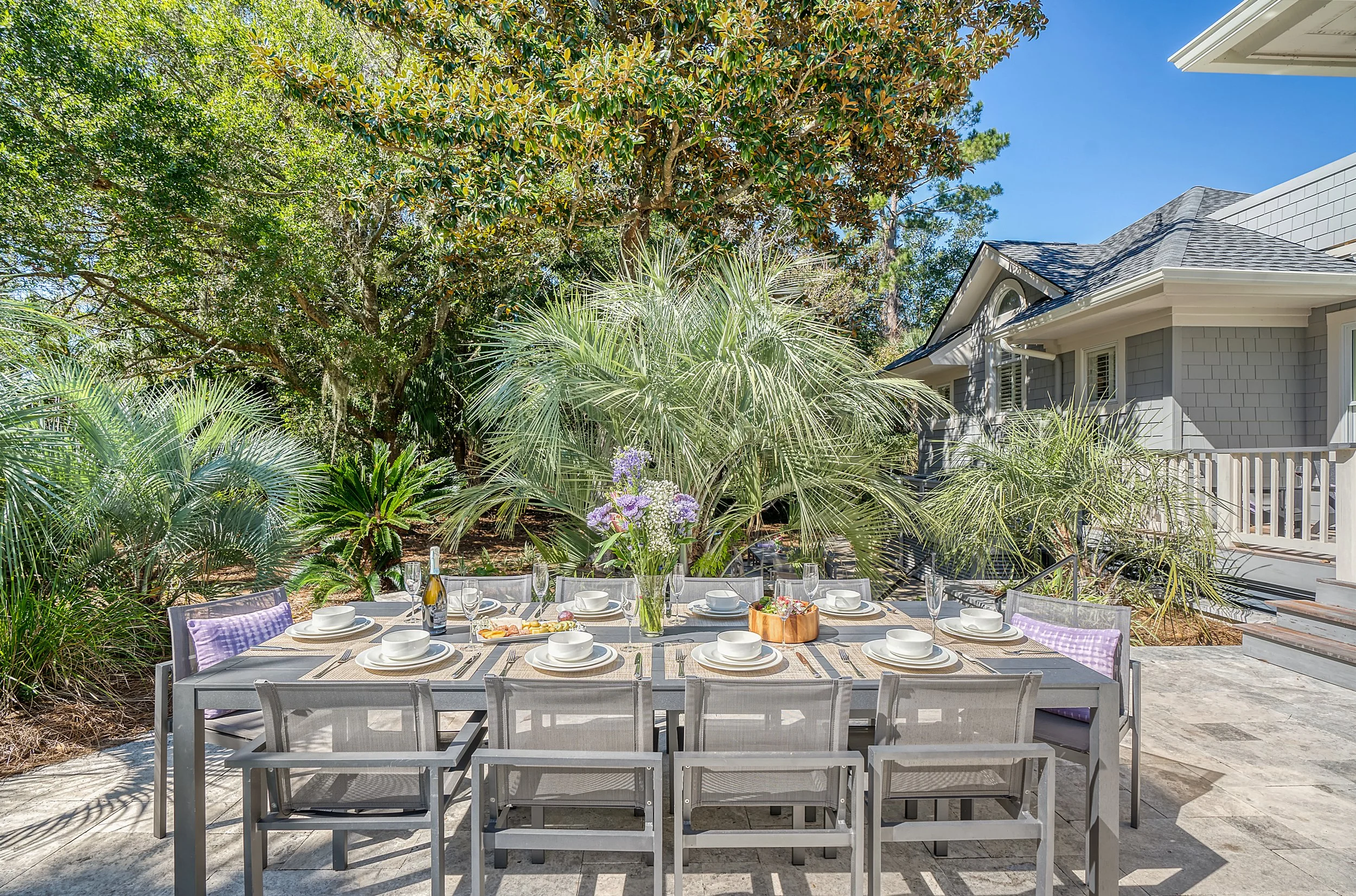 An outdoor dining table set for a meal, surrounded by lush greenery, with a house and clear blue sky in the background.
