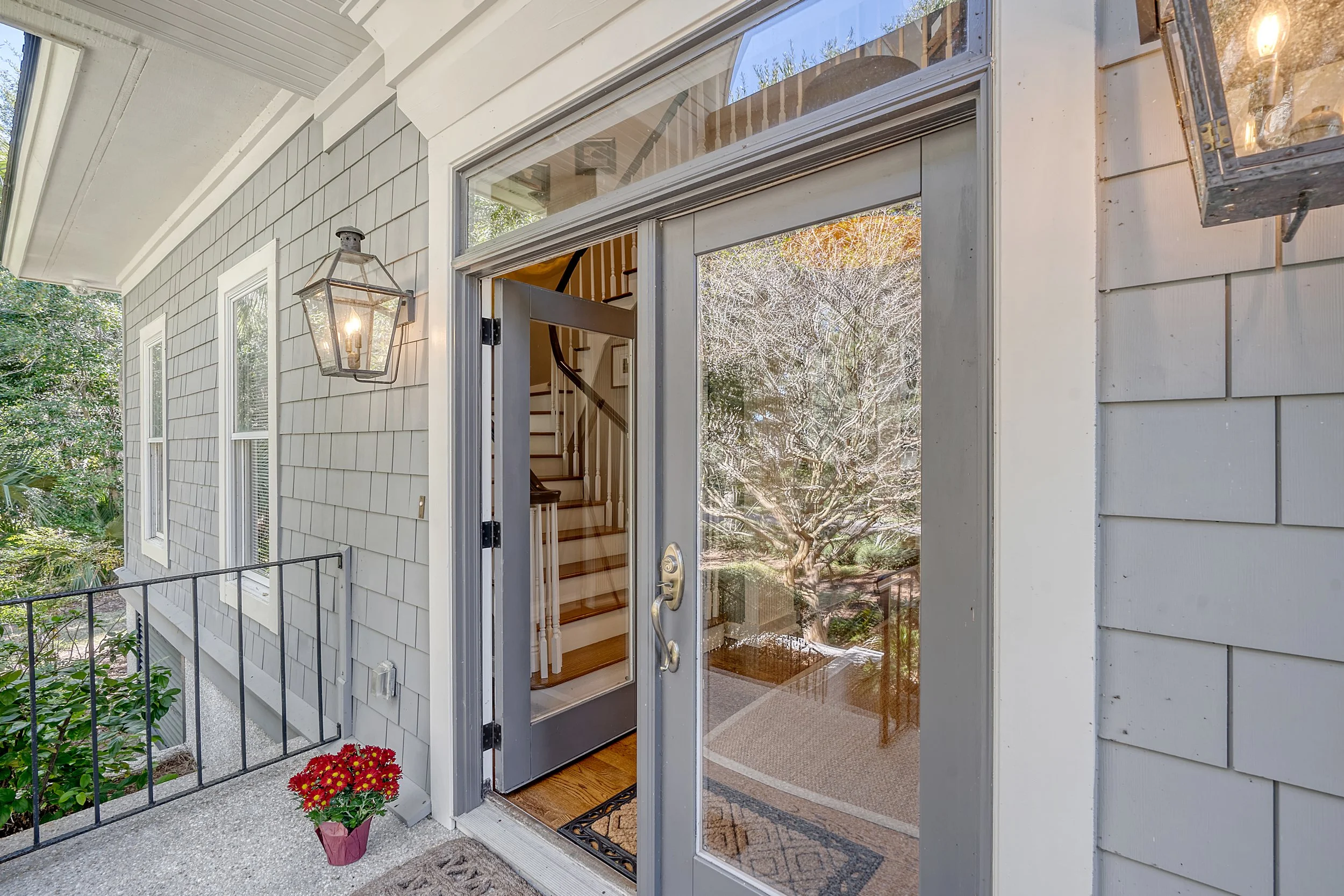 Front door with glass window, open to staircase inside house, outdoor porch with railing and red potted flowers, exterior siding of house, tree visible through glass.