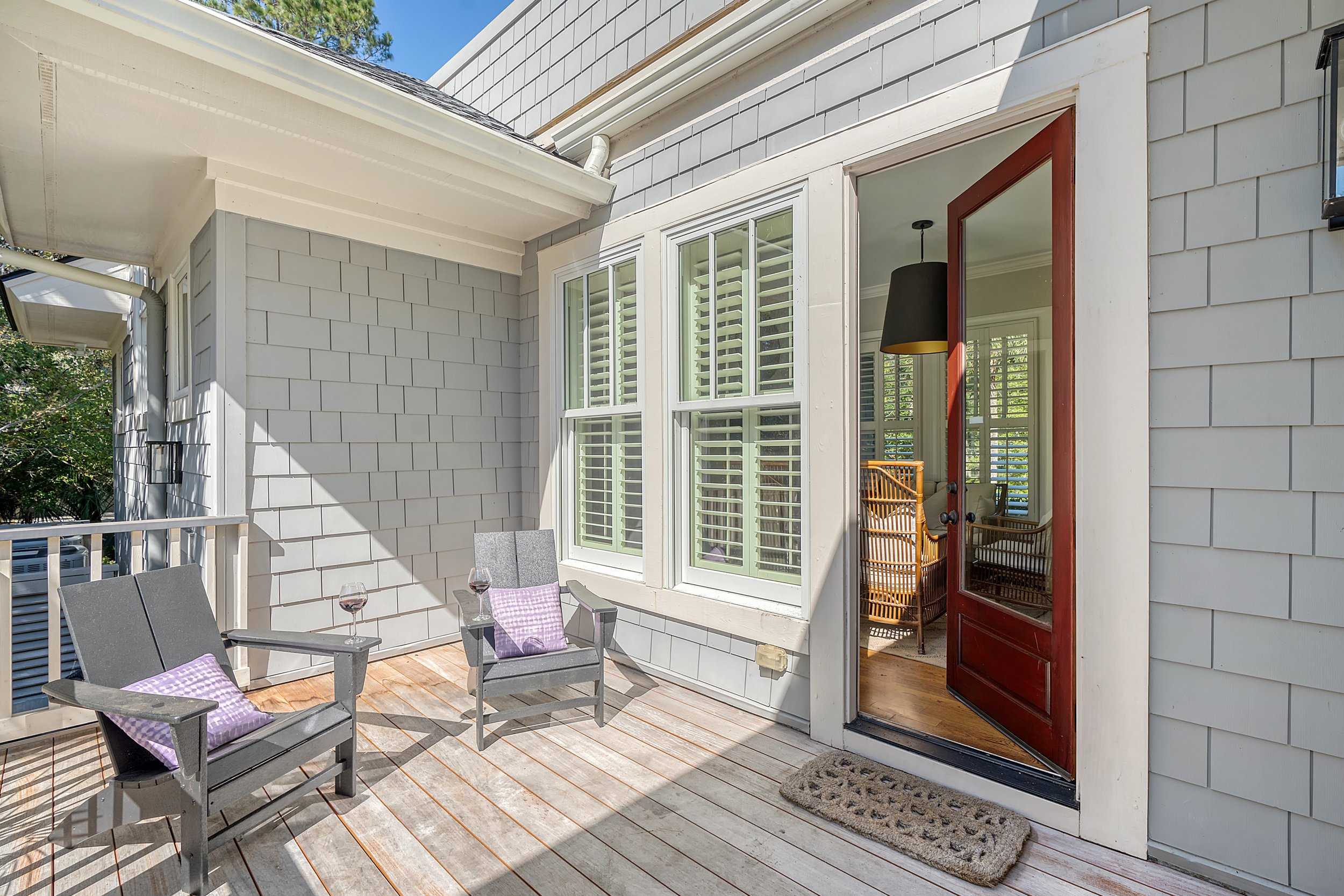 Deck with two gray chairs, each with a purple pillow, and a glass of red wine on each chair's armrest. An open door with a red frame leads inside, showing a black pendant light and wicker chairs inside. The house exterior is painted gray with white t