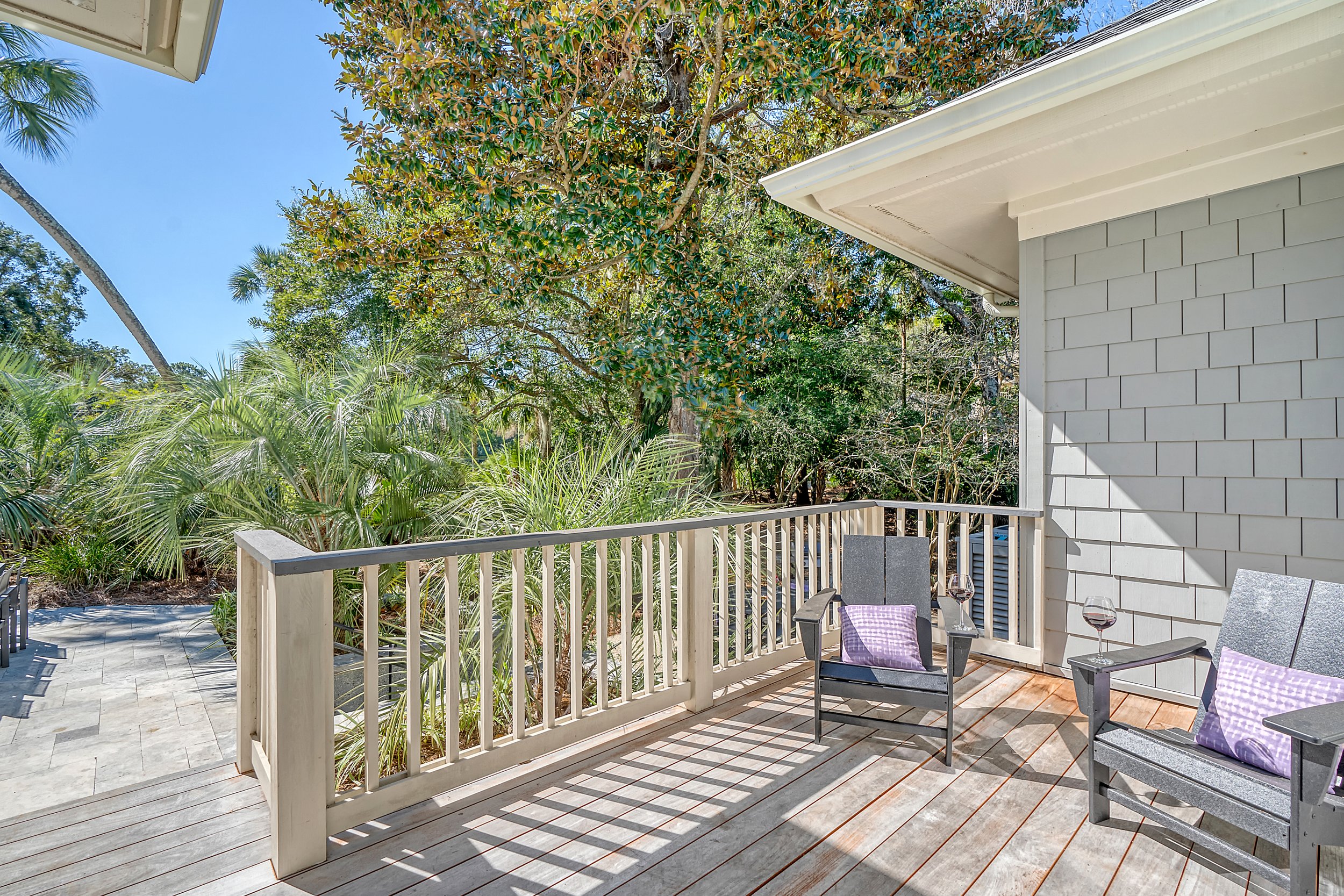 View of a wooden balcony with two chairs holding purple cushions and glasses of red wine, overlooking lush green trees and plants under a clear blue sky.