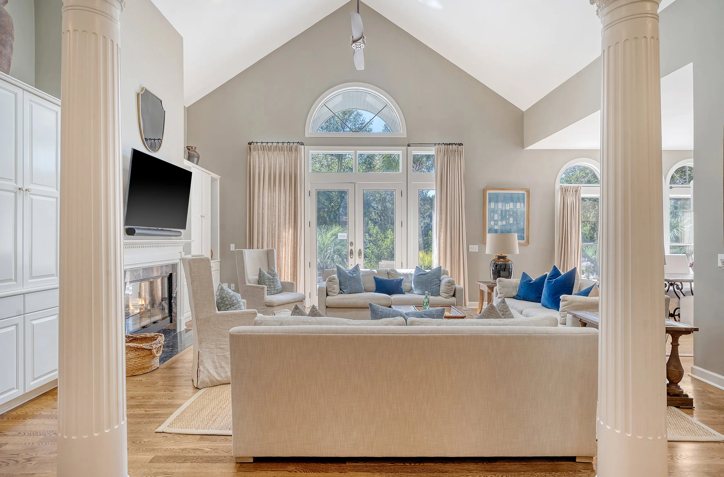 Bright living room with large windows, beige sofas with blue and beige cushions, a fireplace, and a mounted flat-screen TV.