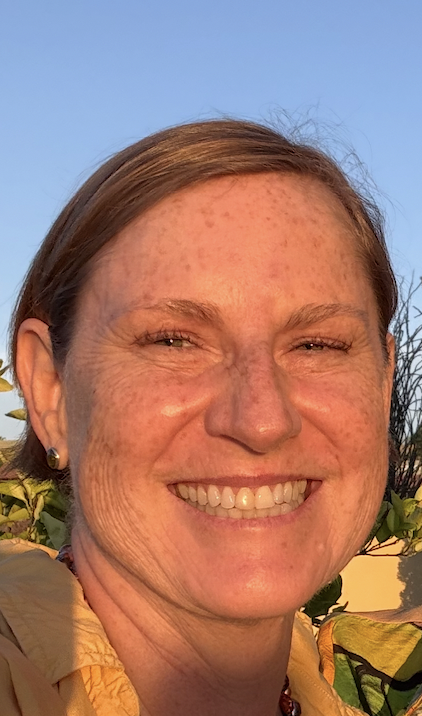 Close-up of a smiling woman with freckles and brown hair outdoors against a clear blue sky.