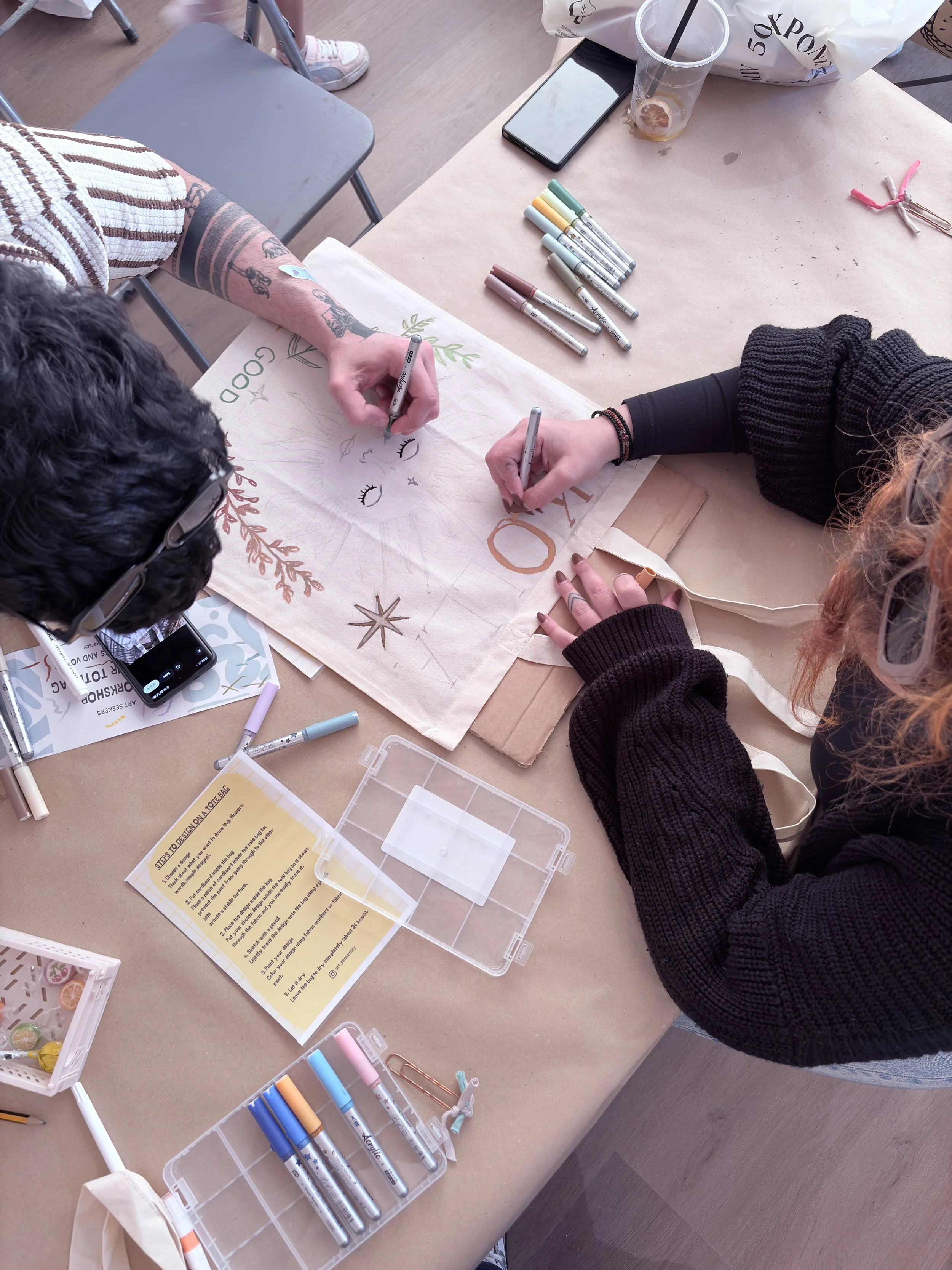 Two people are creating a hand-lettered design on a tote bag with markers. Art supplies, including markers, pens, and decorative elements, are spread across the table. A smartphone, a plastic cup with a drink, and paper instructions are also visible.