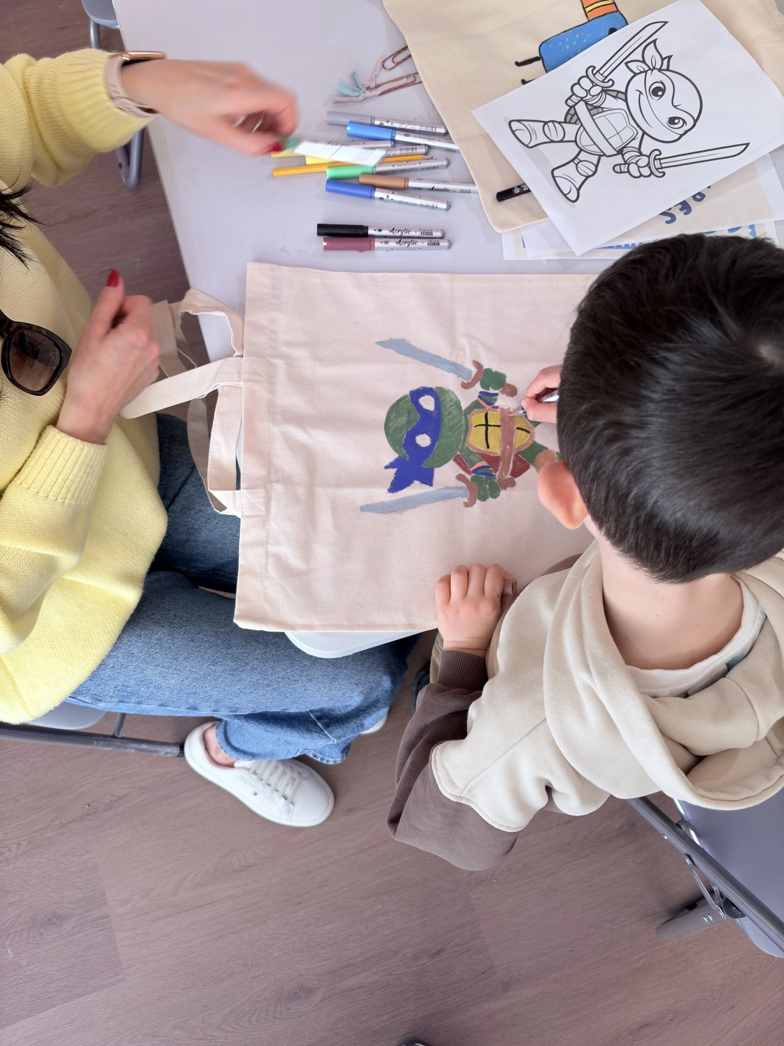 Children coloring a Ninja Turtle drawing on a cloth tote bag at a table with various colored pens and markers.