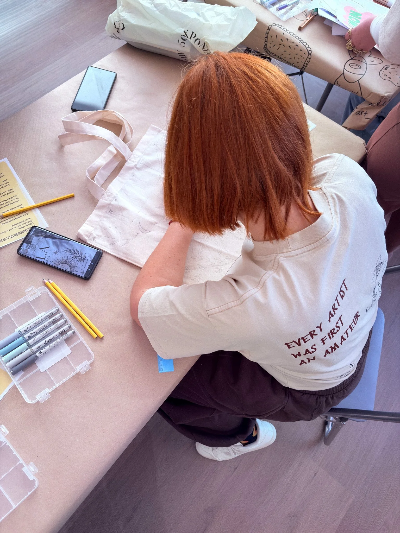 A person with red hair drawing at a desk with art supplies, a smartphone, and a tote bag nearby. The person wears a beige t-shirt with red text on the back.