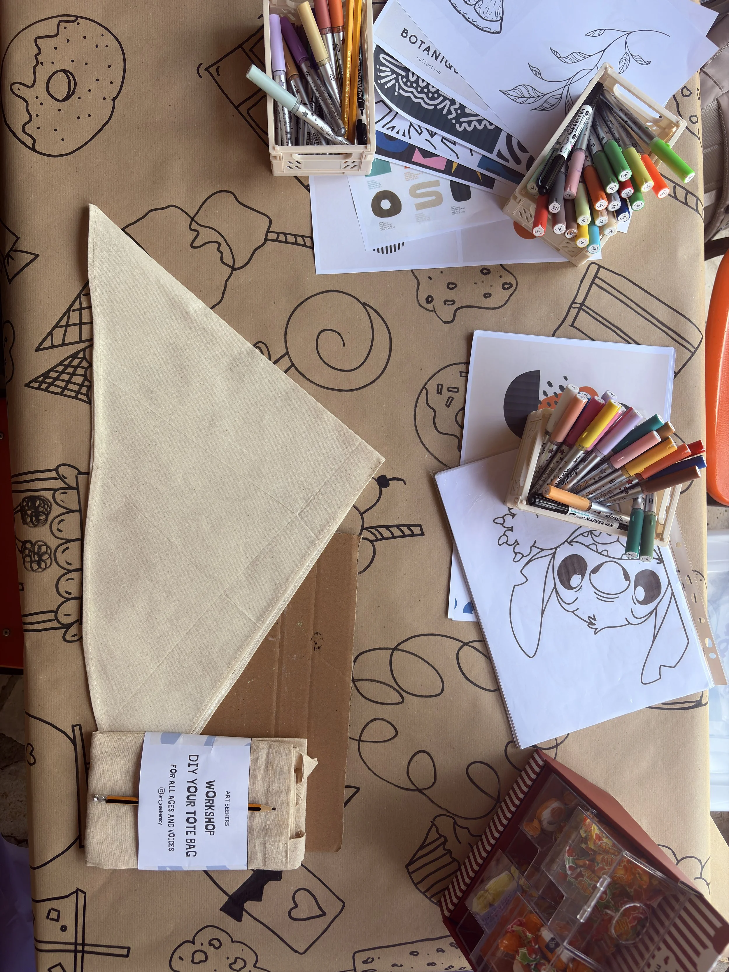 Table covered with a paper featuring line drawings of various food items and desserts, with multiple art supplies including markers in containers, sheets of coloring pages, a beige tote bag with a sign, a box of pencils, and a container of assorted candies.