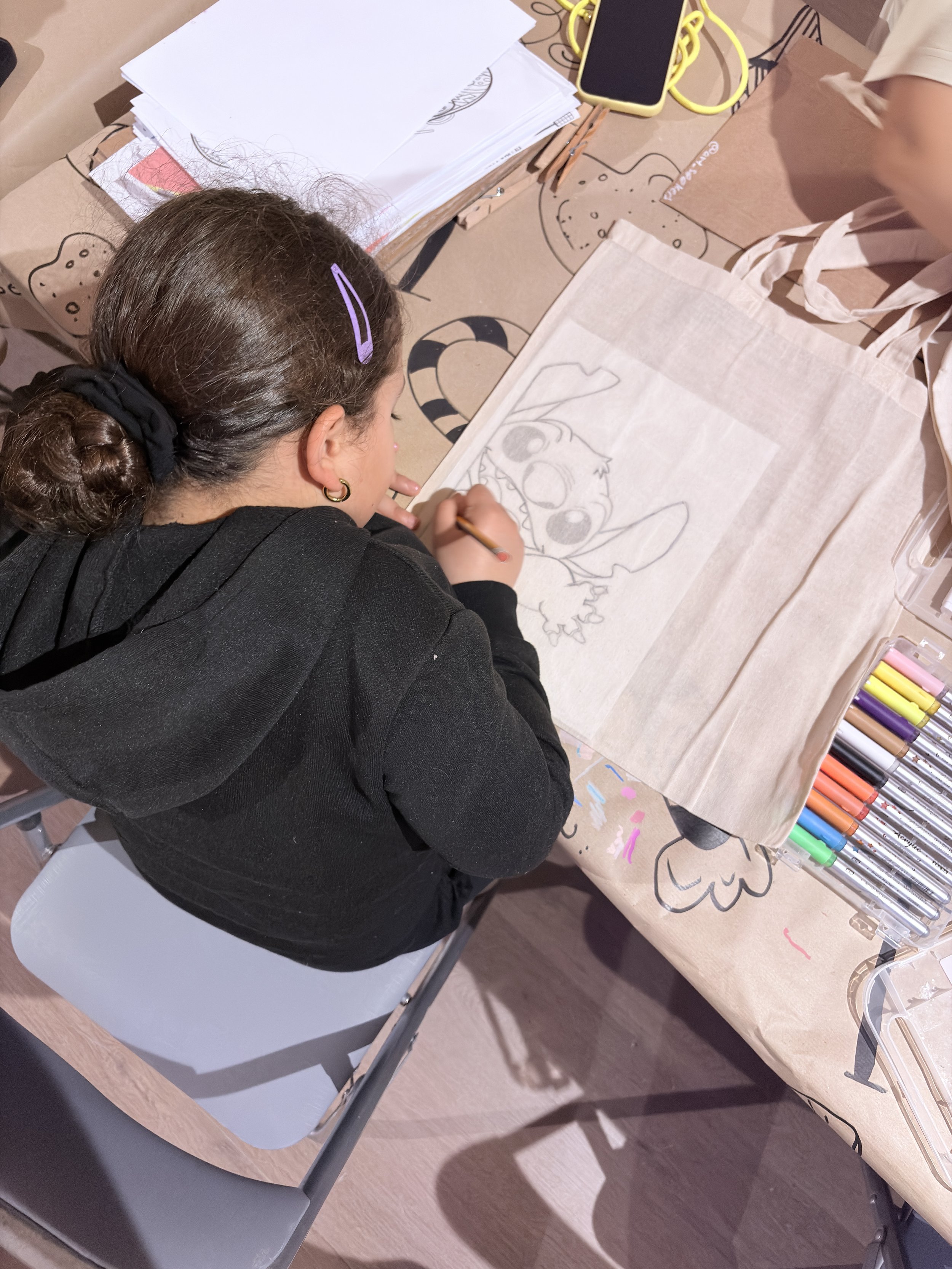 A woman with brown hair tied in a bun, wearing a black hoodie and a purple hair clip, is drawing a cartoon character, possibly a creature with big eyes and ears, on a piece of paper on a table. There are art supplies and papers scattered around.