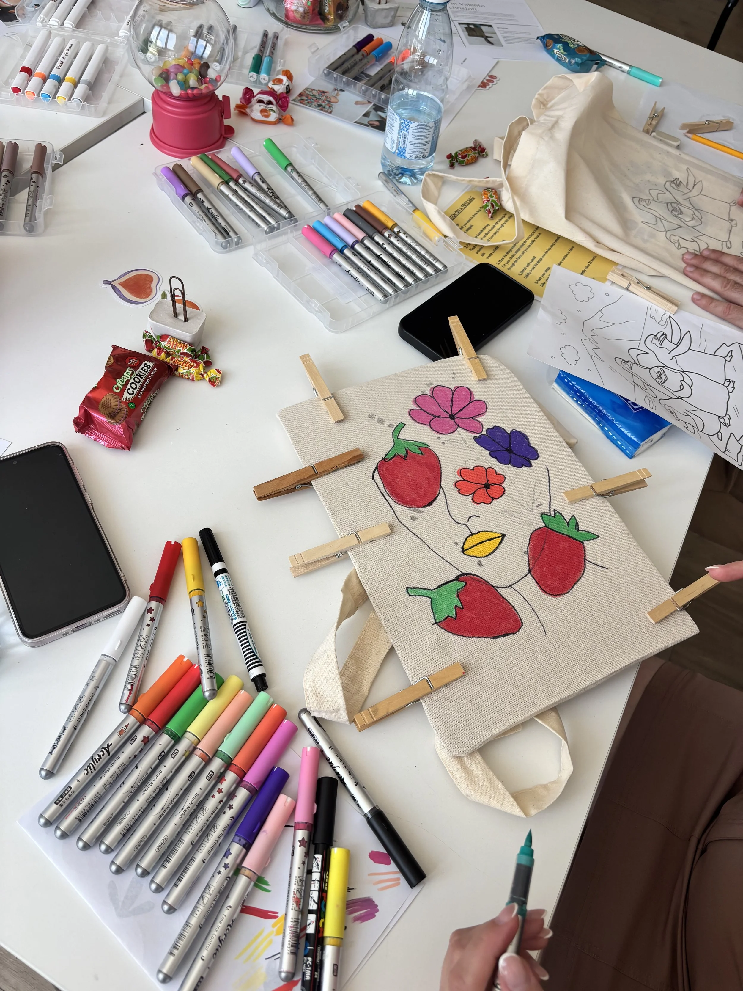 A table with art supplies including markers, a small painting of strawberries and flowers, a water bottle, and a painting tote bag. Someone is holding a marker and working on the tote bag.