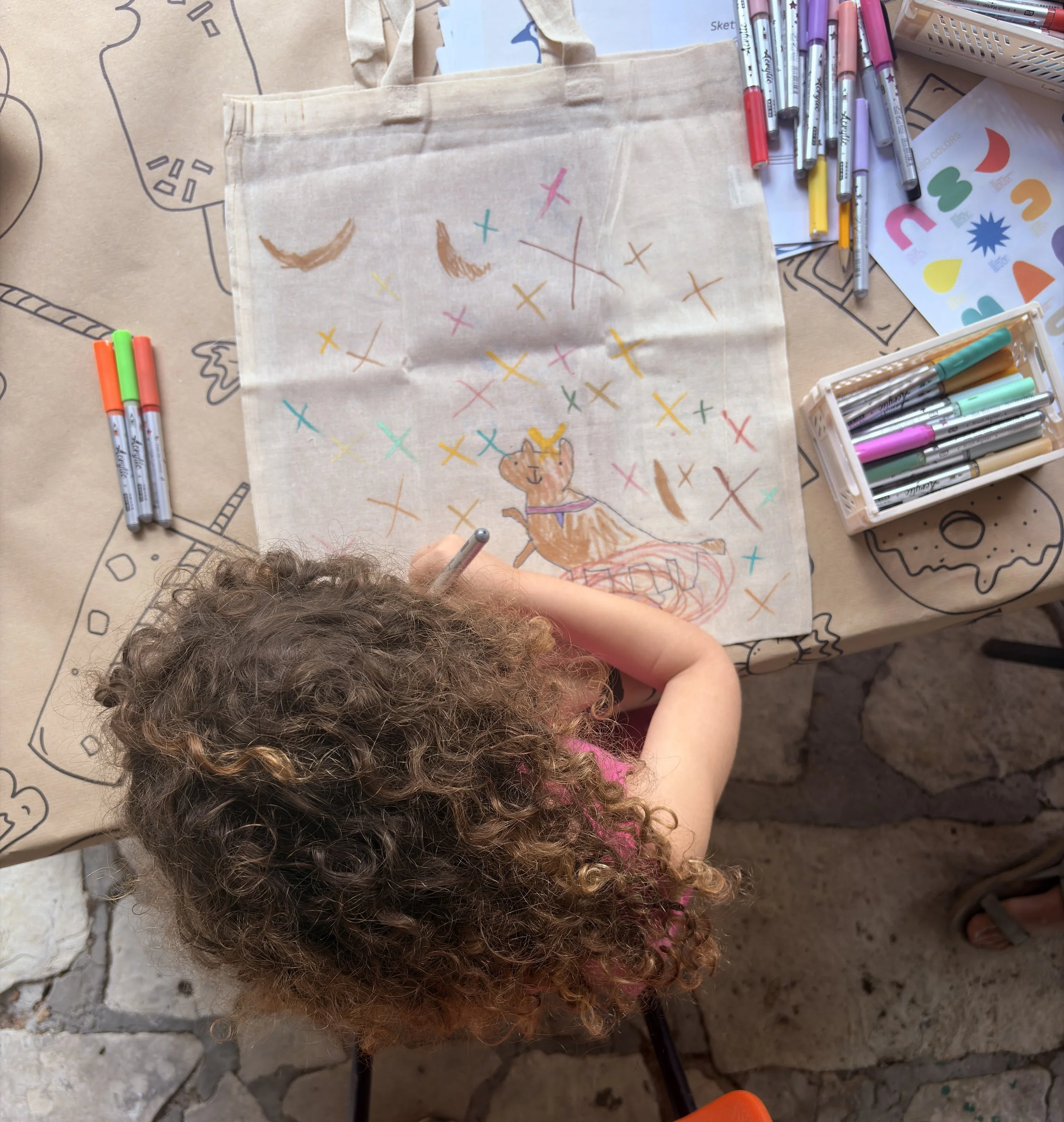 Colorful drawing of a bear sitting on a platform with various X and Y marks and crescent shapes around it, surrounded by markers and drawing supplies on a table.
