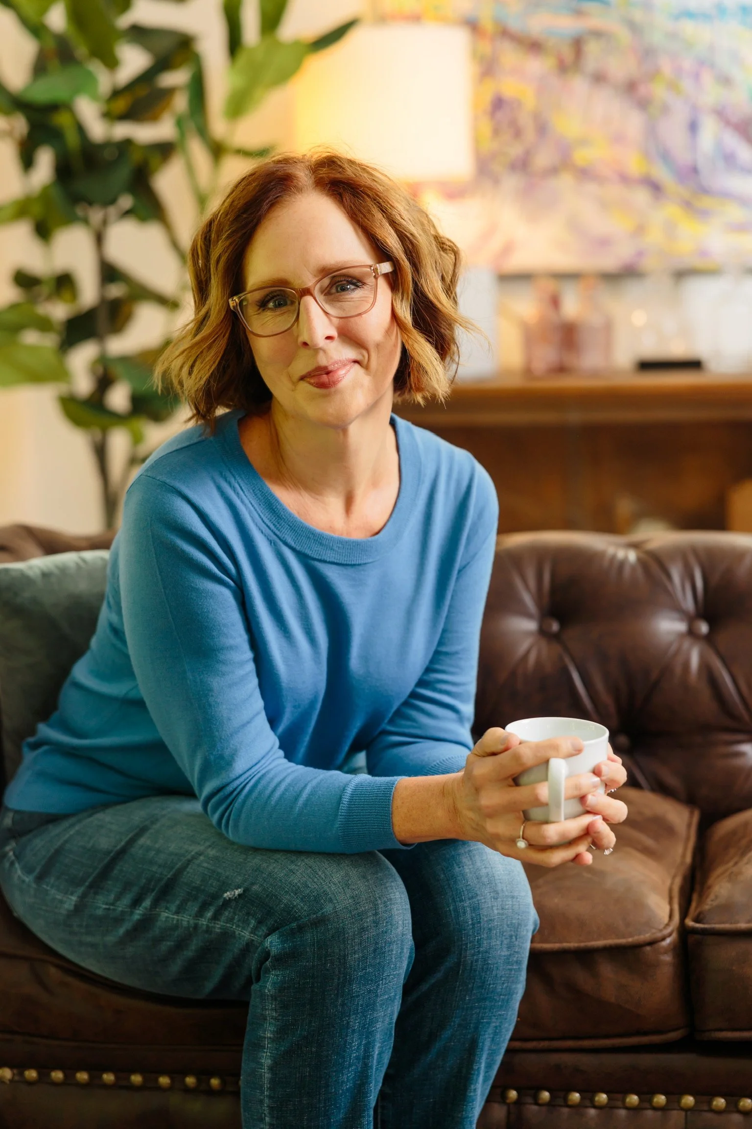 A woman with glasses and short, wavy, red hair sitting on a brown leather couch, holding a white mug, in a cozy living room.