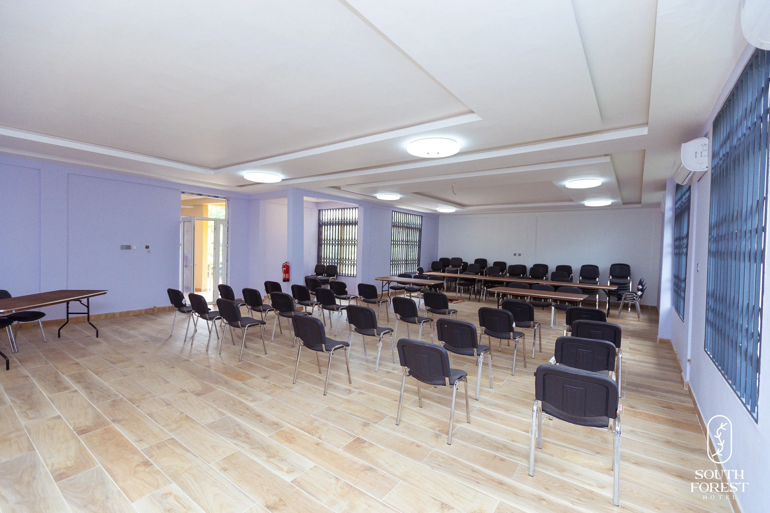 Empty conference room with rows of black chairs, some tables at the front, light wood flooring, and large windows with vertical blinds.
