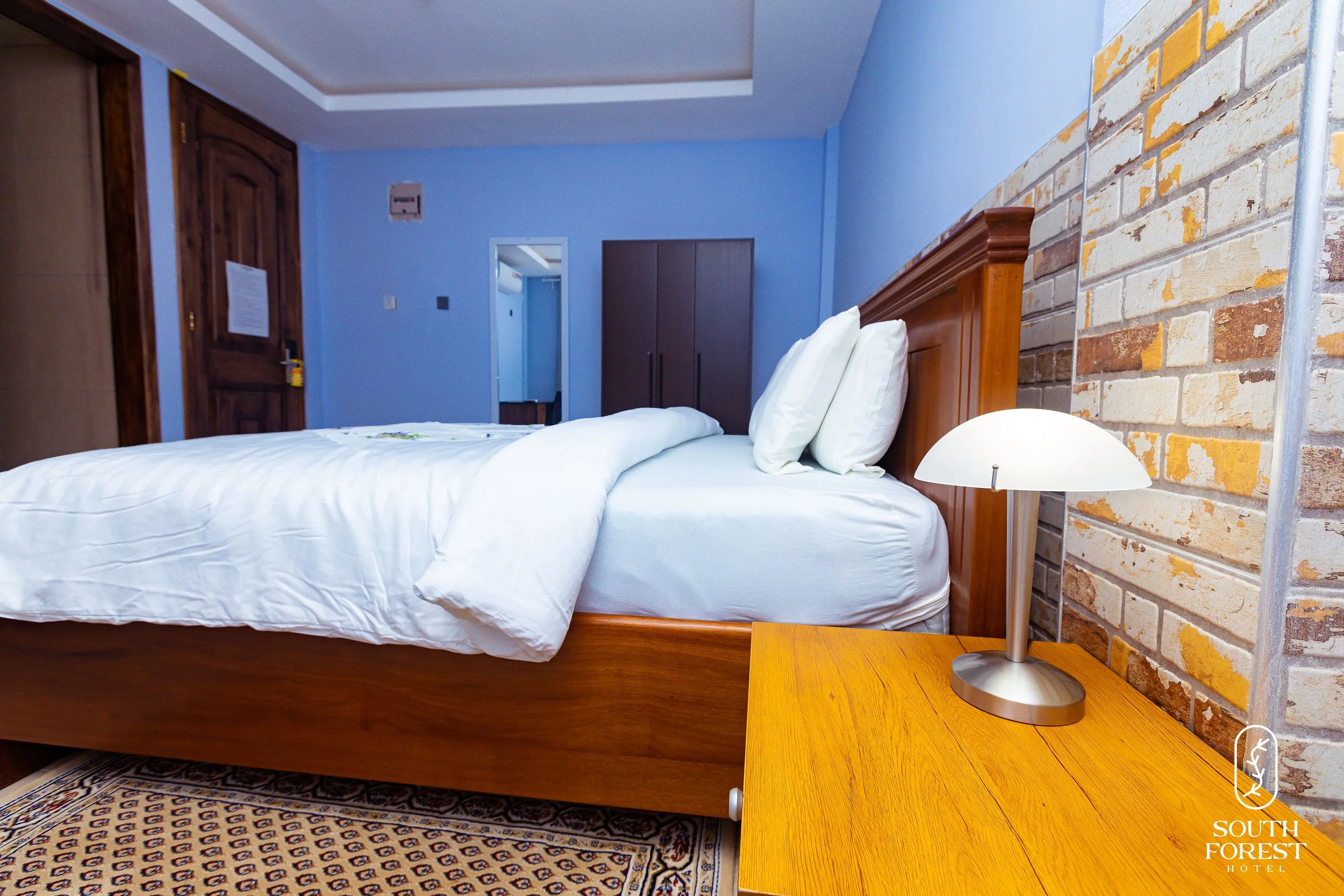 A hotel room with a bed, white bedding, and pillows, a wooden nightstand with a lamp, a brick accent wall, and blue painted walls.