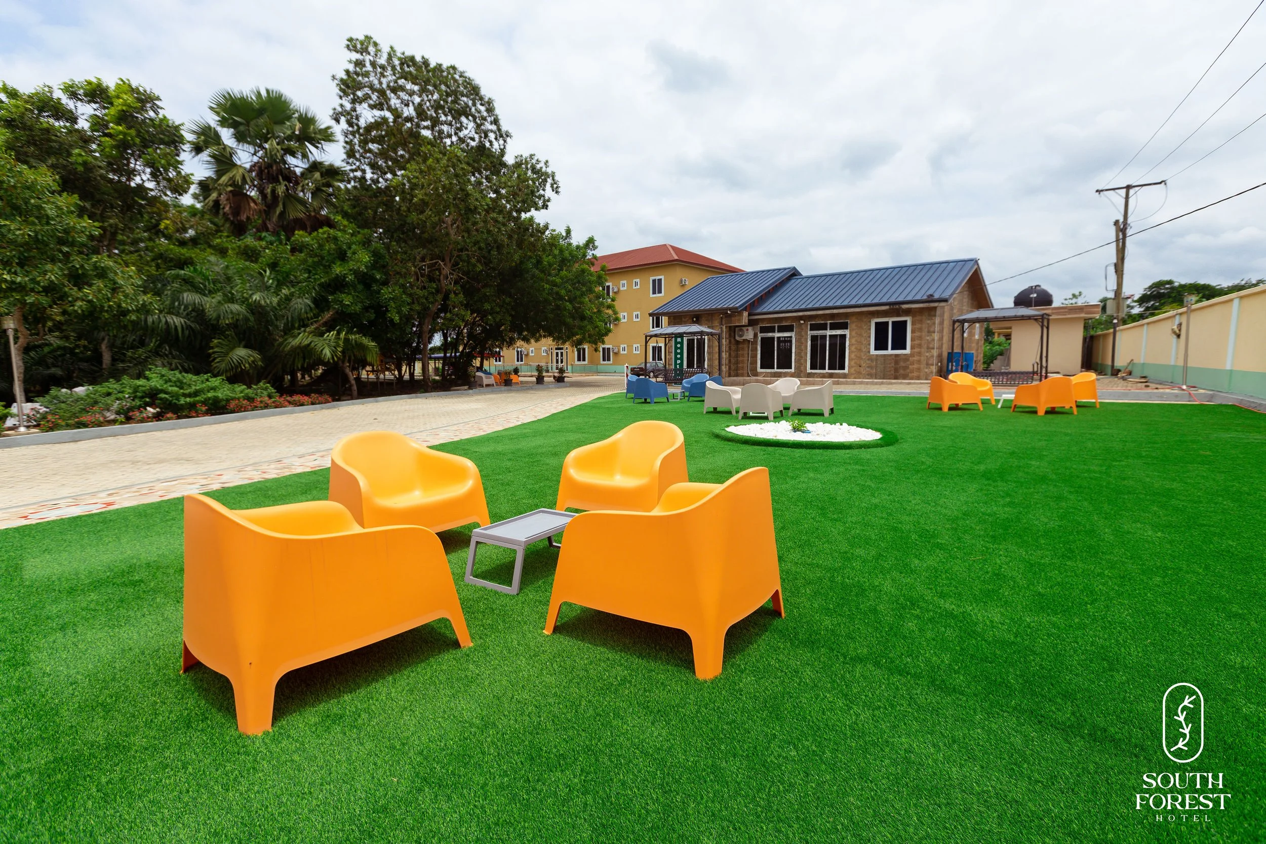 Outdoor seating area at South Forest Hotel with orange chairs, colorful chairs, and a small table on a green lawn, with trees and a building in the background