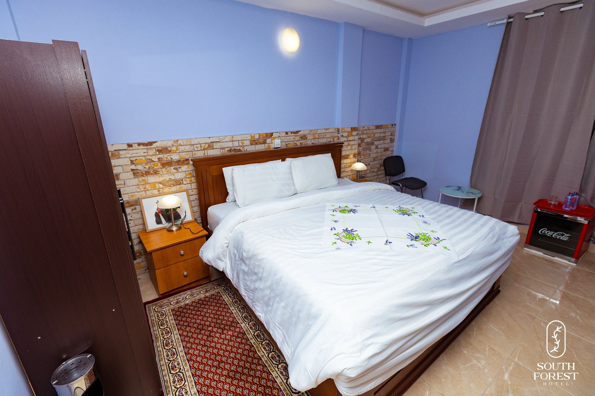 Hotel room with a bed, nightstands, lamps, a chair, a mini fridge, and curtains. The wall behind the bed has a brick accent, and the room is decorated in warm and cool tones.