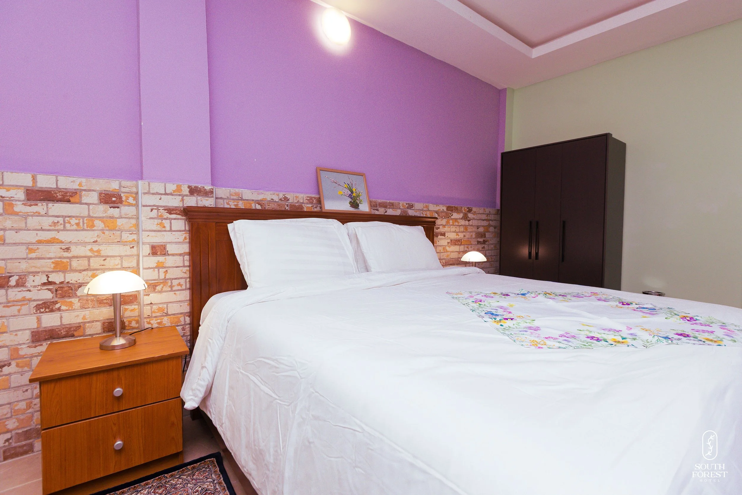 A hotel bedroom with a white bed, purple and brick wall background, wooden headboard, bedside table, lamps, framed artwork, and a black wardrobe.