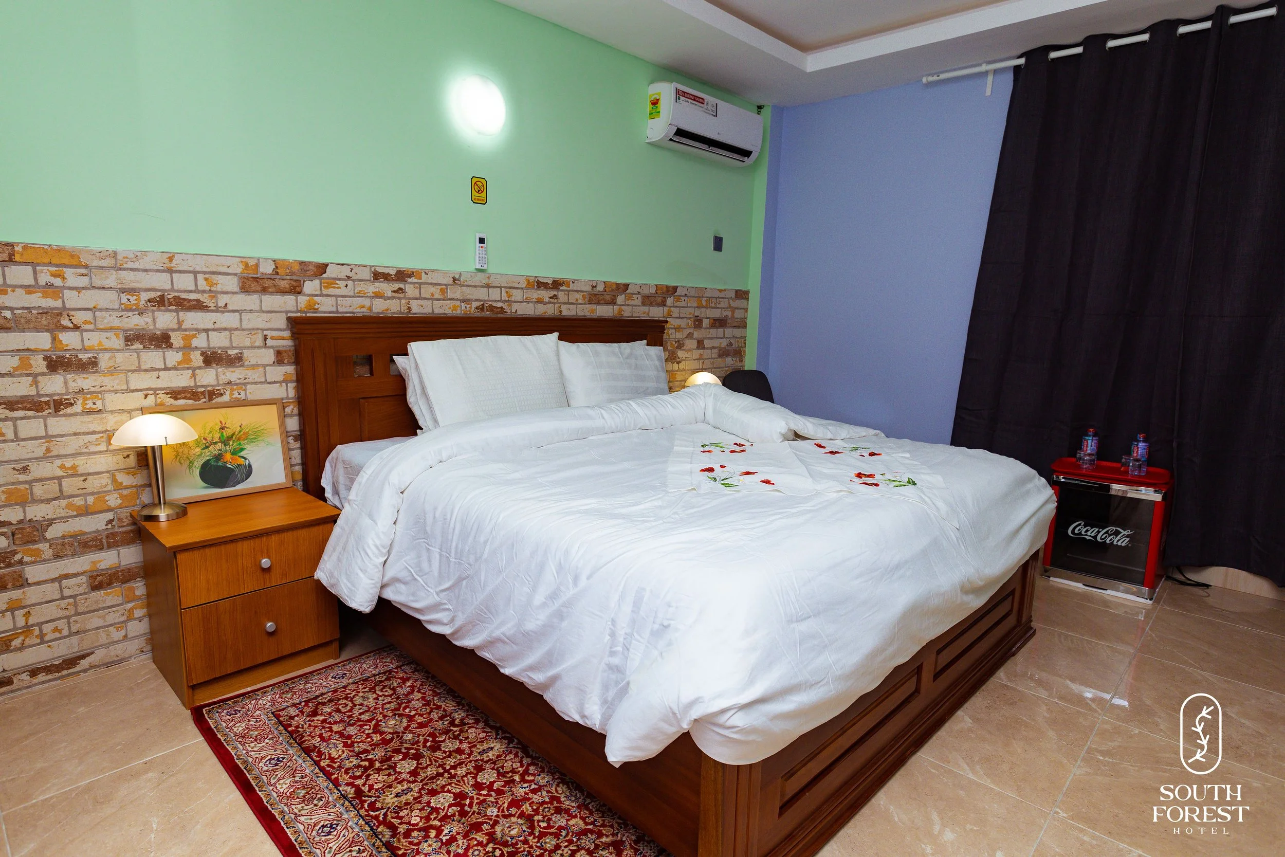 A hotel room with a king-sized bed, white linens, a wooden headboard, and pillows, next to a small wooden nightstand with a lamp. There is a red Coca-Cola mini fridge in the corner, black curtains, and an air conditioning unit on a wall painted dual 