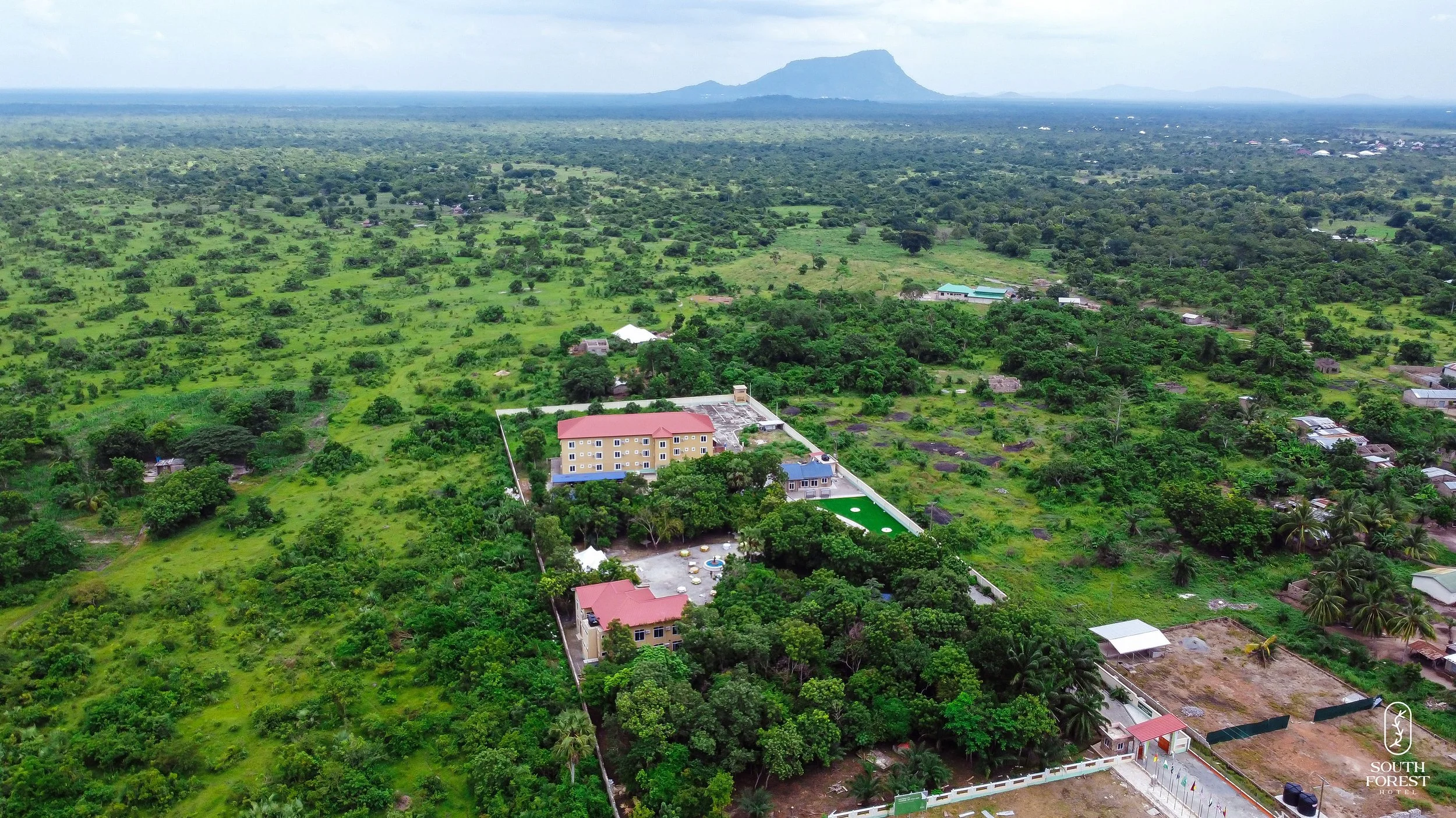Aerial view of lush green landscape with a hotel complex, surrounded by trees, with mountains in the distance.