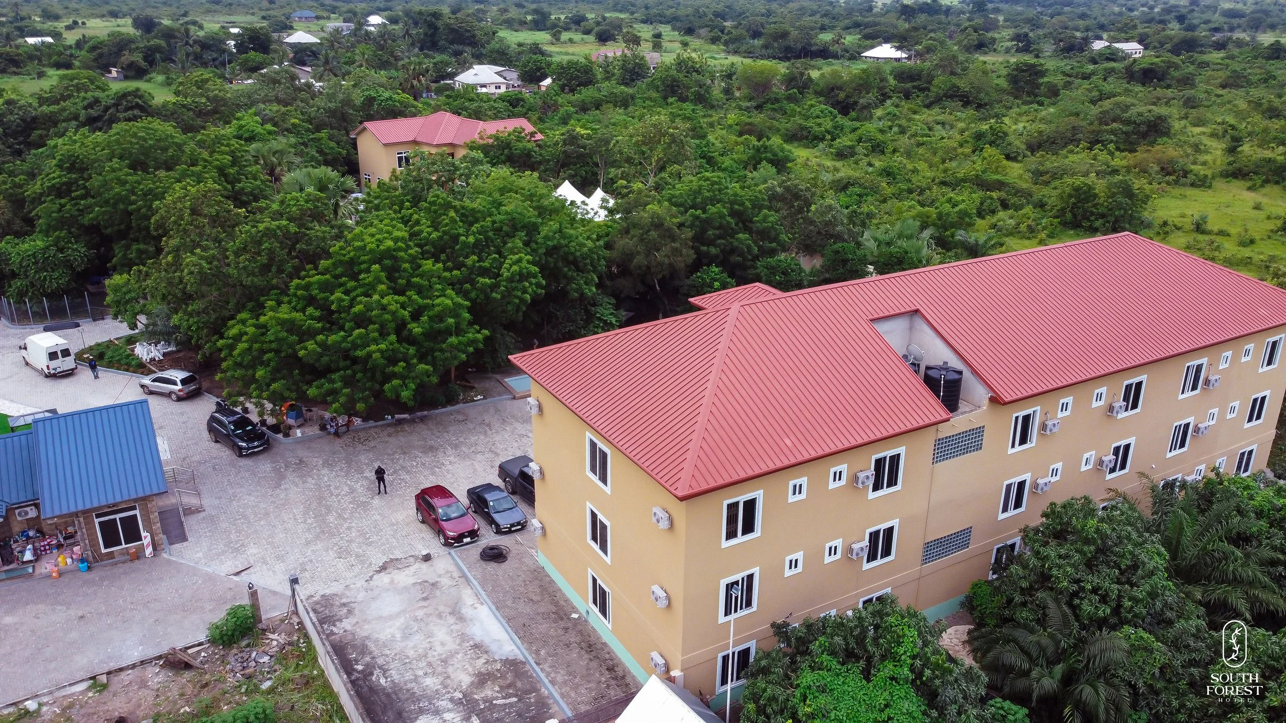 Aerial view of a residential area with a large yellow apartment building with a red roof, surrounded by green trees and other houses, some with white rooftops. Vehicles are parked outside the building, and there are a few people visible near the park
