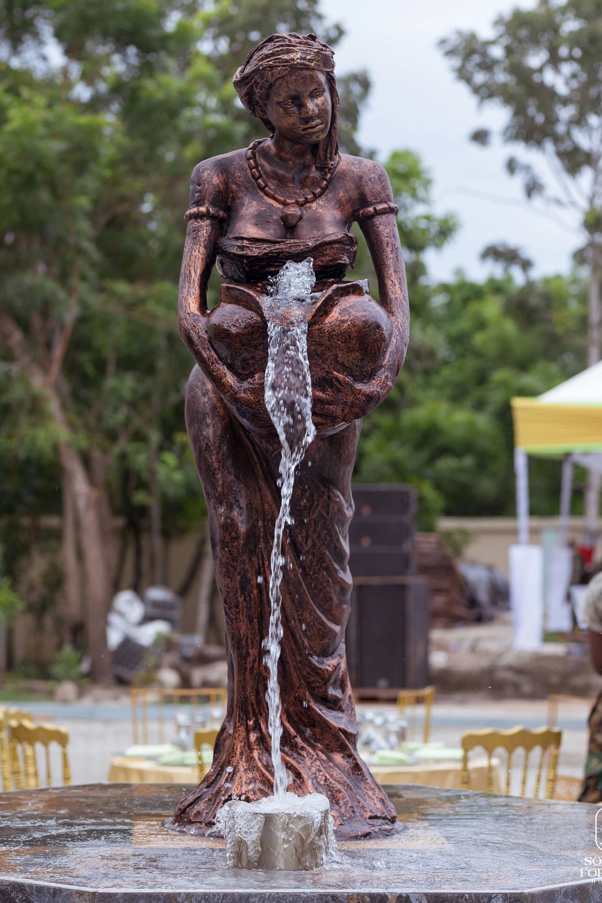 Bronze statue of a woman with water flowing from her lap, outdoors with trees in the background.