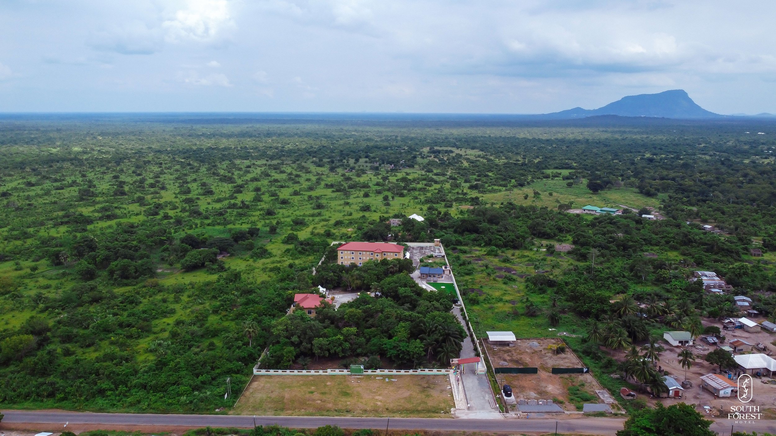 Aerial view of lush green landscape with a hotel complex surrounded by trees, a road in the foreground, and mountains in the distance under a partly cloudy sky.